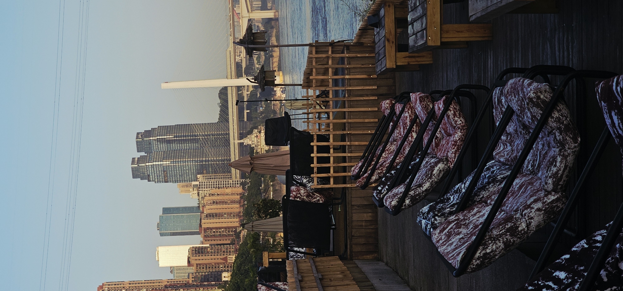 The image shows an outdoor seating area with several lounge chairs and tables on a wooden deck. In the background, a city skyline with modern skyscrapers and a bridge is visible across a body of water. The sky is clear and light blue. The lounge chairs have black frames and cushions with a marbled pattern of red, white, and black. Two wooden tables are also present. Several closed umbrellas and some covered chairs are located on a higher level of the deck behind a wooden fence. The lighting suggests it is either early morning or late afternoon. The scene appears to be located in Nan'an District, China, based on the distinct architecture visible in the skyline, particularly the pair of prominent twin skyscrapers and the bridge design.