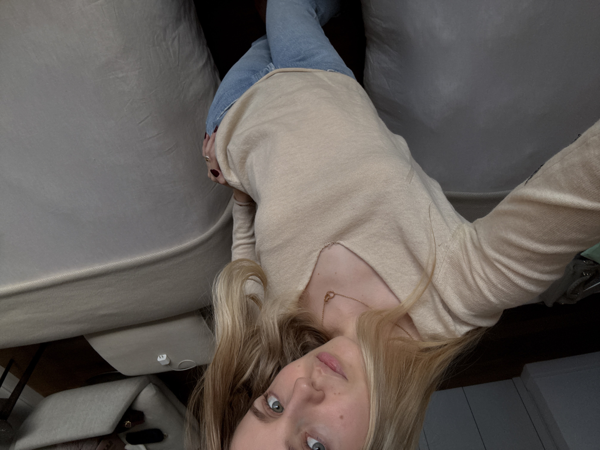 A young woman is pictured from an overhead perspective. She has fair skin, light blond hair, and blue eyes. She is wearing a beige, long-sleeved top and blue jeans. A thin gold necklace is visible around her neck. She appears to be lying down, partially wedged between a couch and what looks like another piece of furniture. The setting appears to be indoors, with elements suggesting a domestic environment. The location is not discernible from the visual information provided.