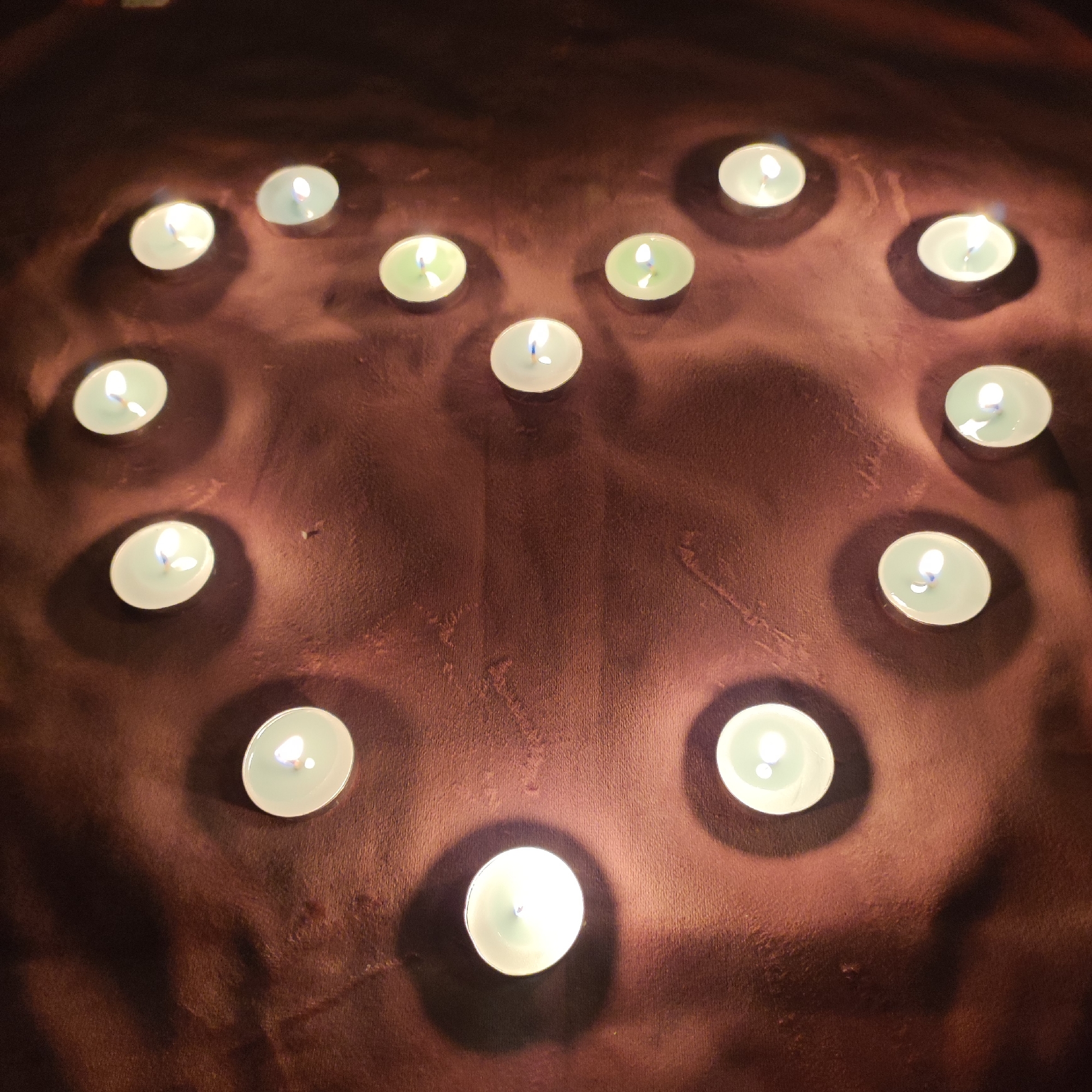 The image shows a dark brown, textured surface, likely fabric, with fourteen lit tea light candles arranged in a rough heart shape. The candles have small, white, cylindrical housings and emit a warm, yellow-white light. Their flames are visible, some flickering. The lighting creates soft shadows and highlights on the fabric. There are no people or other objects visible in the frame. The location in Kineshma, Russia, is not discernible from the image content.