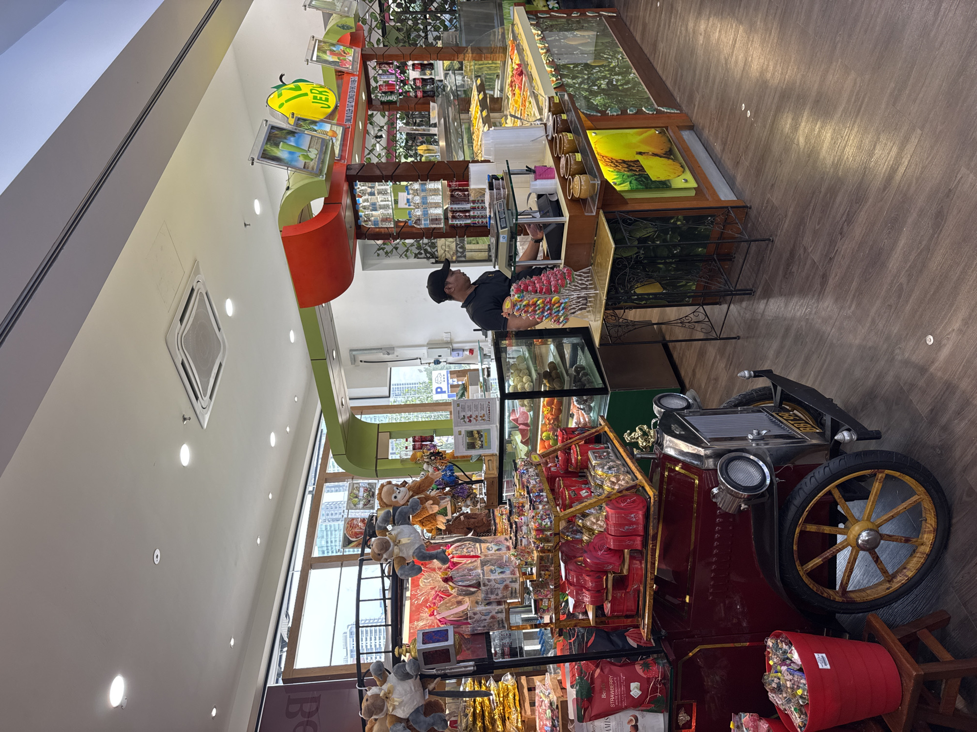 The image shows the interior of a candy store located in Kuala Lumpur, Malaysia. The focal point is a vintage-style car converted into a display stand stocked with various candies, chocolates, and stuffed animals. A sales clerk wearing a black uniform and cap stands behind a counter offering a selection of lollipops and sweets. The background features shelves with more candies and drinks. The store is brightly lit.