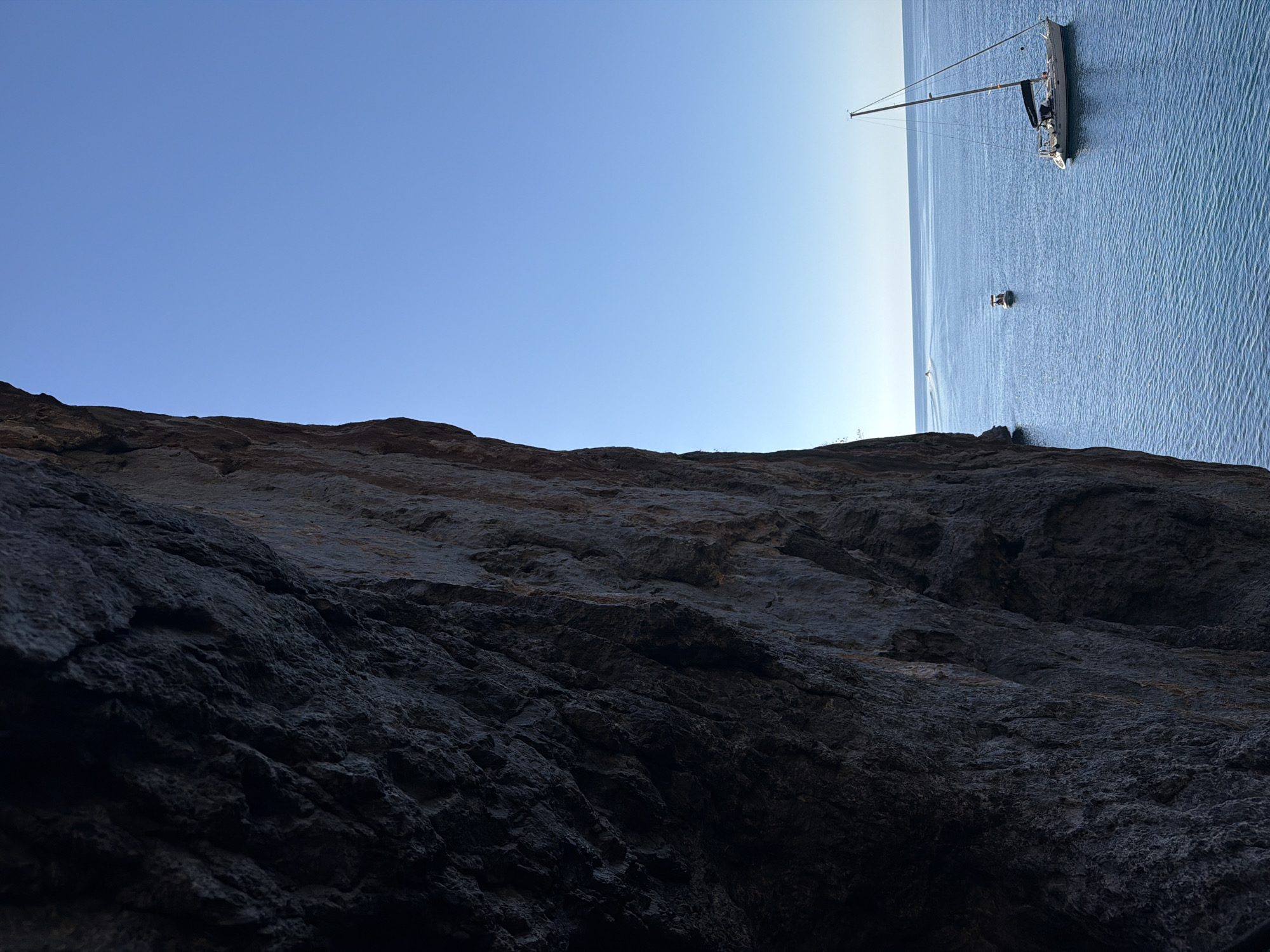 A tilted, low-angle shot depicts a craggy, dark rock face in the foreground, occupying the bottom half of the frame. Above the rocks is a clear blue sky transitioning into a view of the ocean, where a sailboat and a small motorboat are visible. The location is Escorca, Spain, based on the landscape and coastal features.