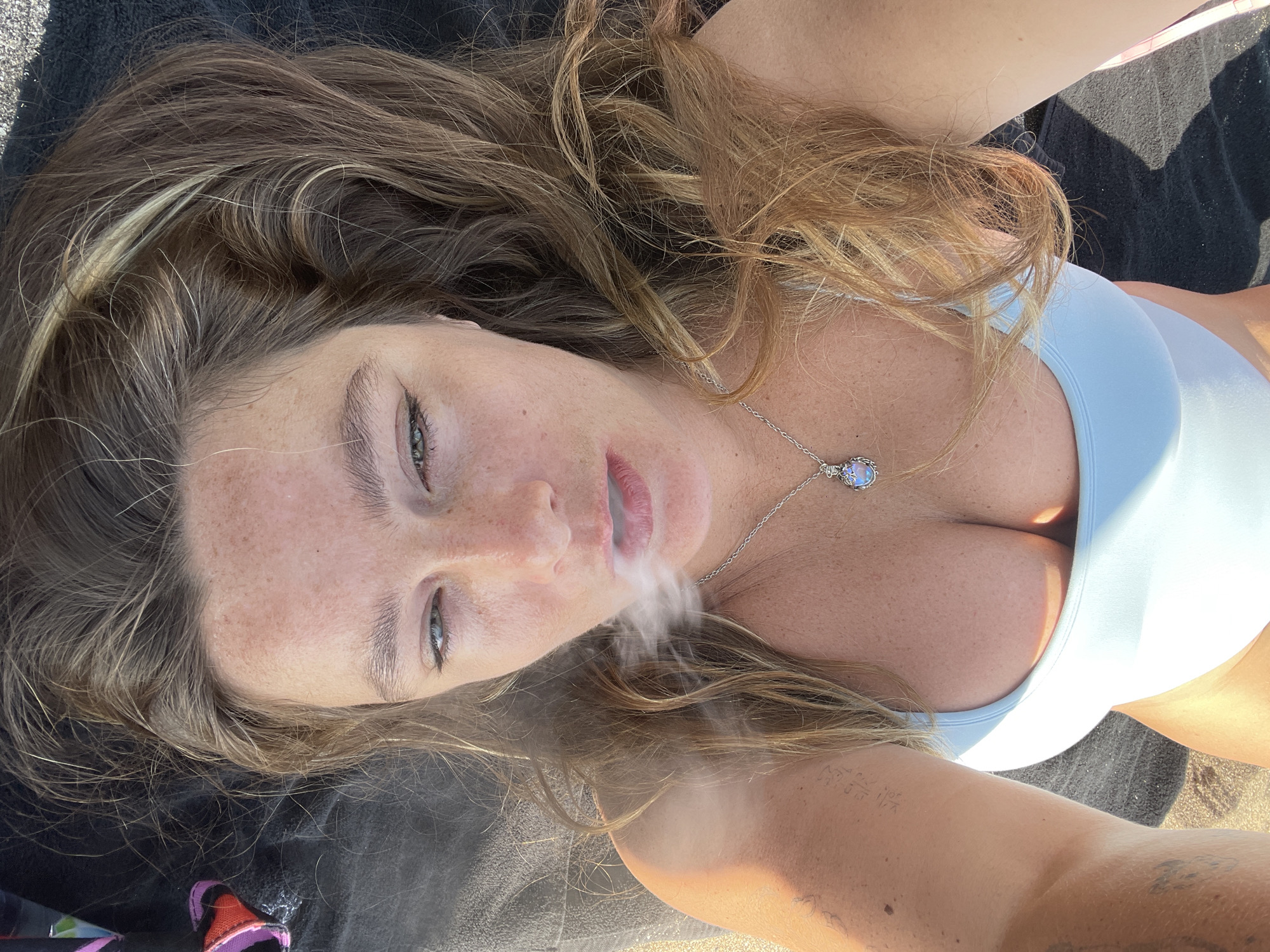 This is a high-angle, close-up, outdoor, POV shot of a young woman lying down on a dark-colored surface, possibly a towel or blanket on sand, as suggested by the texture and scattered dark specks. She has long, wavy, light brown hair with blonde highlights, and her face is speckled with freckles. She is wearing a light blue bikini top and a silver necklace with a pale blue pendant. Her eyes are open, and she appears to be exhaling a cloud of smoke or vapor. The lighting suggests it is daytime, likely sunny, casting shadows and highlighting her skin. The overall mood is relaxed and sensual, with the focus on her facial expression and the visible cleavage. There are no discernible text elements in the image.