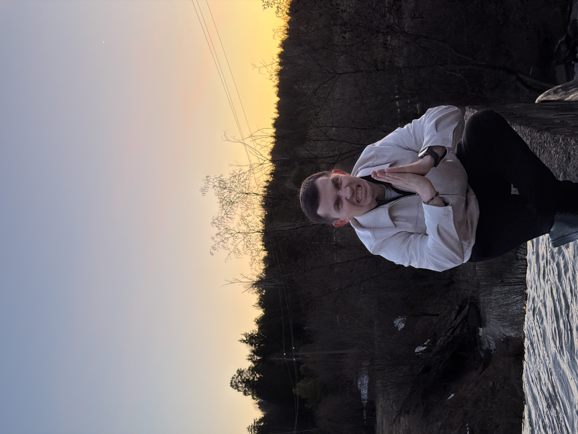 The image, taken in Kamennoye, Russia, captures an outdoor scene during either sunrise or sunset. The sky displays a striking gradient, transitioning from a light blue at the top left to warm orange and yellow hues near the horizon on the right. A small, faint white dot, possibly a star or planet, is visible in the upper left sky.
The scene features a young man positioned on a dark, rocky or stone bank next to a body of water, possibly a river or lake, which shows distinct ripples on its surface. He is wearing a light-colored, long-sleeved jacket or shirt and dark trousers. His hands are clasped together in front of him, fingers interlocked, reminiscent of a prayer or bowing gesture, but with an intense, animated facial expression. His mouth is open in what appears to be a shout, yell, or an exaggerated, powerful grimace, with furrowed eyebrows. He has short, dark hair, a watch on his left wrist, and a small, beaded bracelet on his right wrist.
Behind the man and across the horizon, a dense, dark forest of mostly bare trees is visible, suggesting a colder season like winter or early spring. Several power lines stretch horizontally across the sky above the tree line. The overall lighting is dim, characteristic of twilight. No visible text is present in the image.