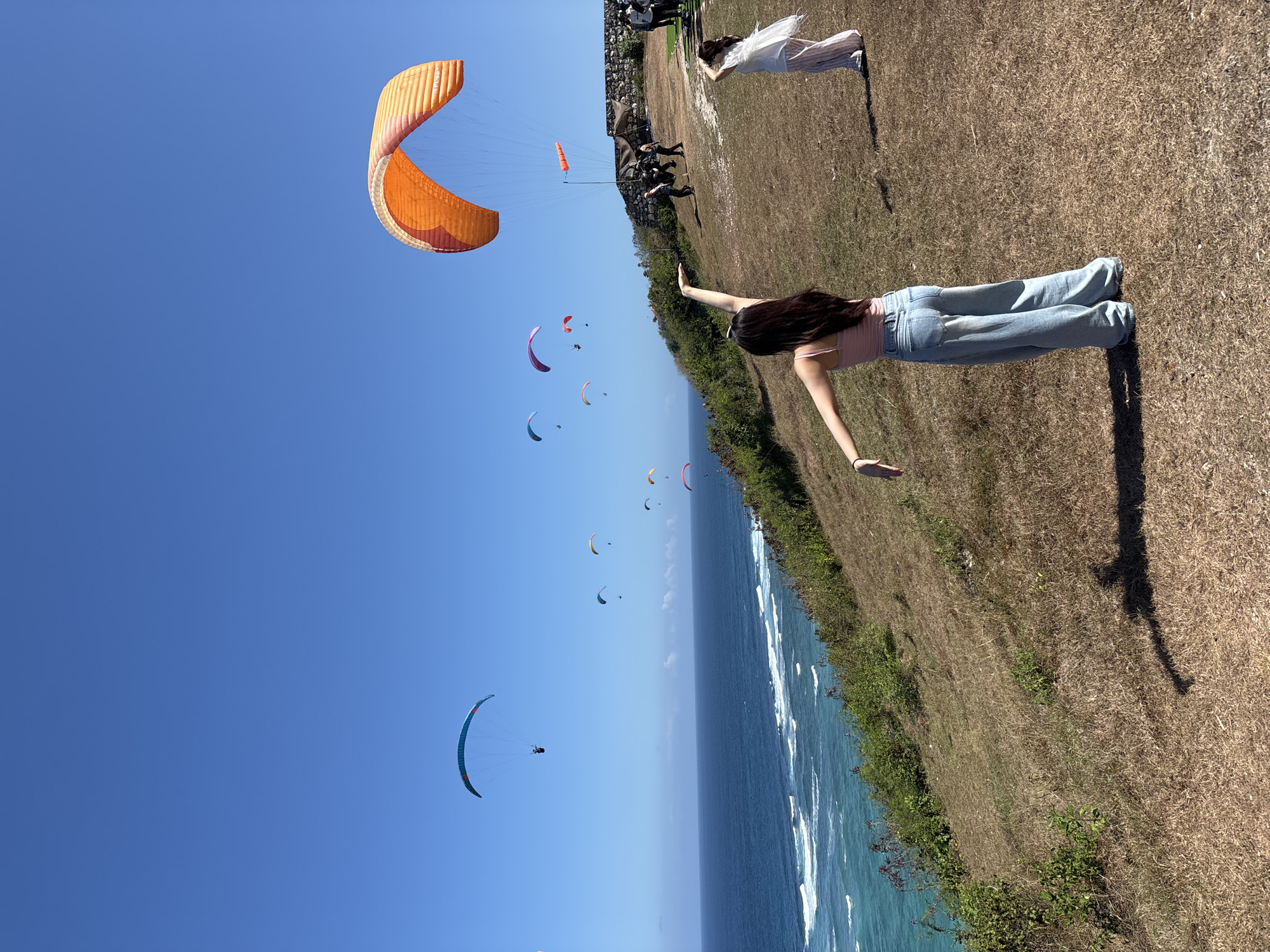 The image depicts a scenic coastal location in Kuta Selatan, Indonesia, where paragliders are active. The foreground features a grassy hillside with two young women facing away from the camera, enjoying the view. The woman on the left is wearing a pink tank top and light blue wide-leg jeans, with her arms outstretched. The woman on the right is wearing a white top and striped pants, holding her hand up to her face. Several paragliders, varying in color and size, are visible in the bright blue sky, soaring above the turquoise ocean. The ocean exhibits white waves crashing near the shore. In the middle ground, a group of people are gathered near a stone wall, possibly preparing for paragliding or observing the activity. The terrain is dry and covered in brown grass, with some green shrubbery on the slope leading down to the sea. The overall scene conveys a sense of adventure and natural beauty on a clear, sunny day.