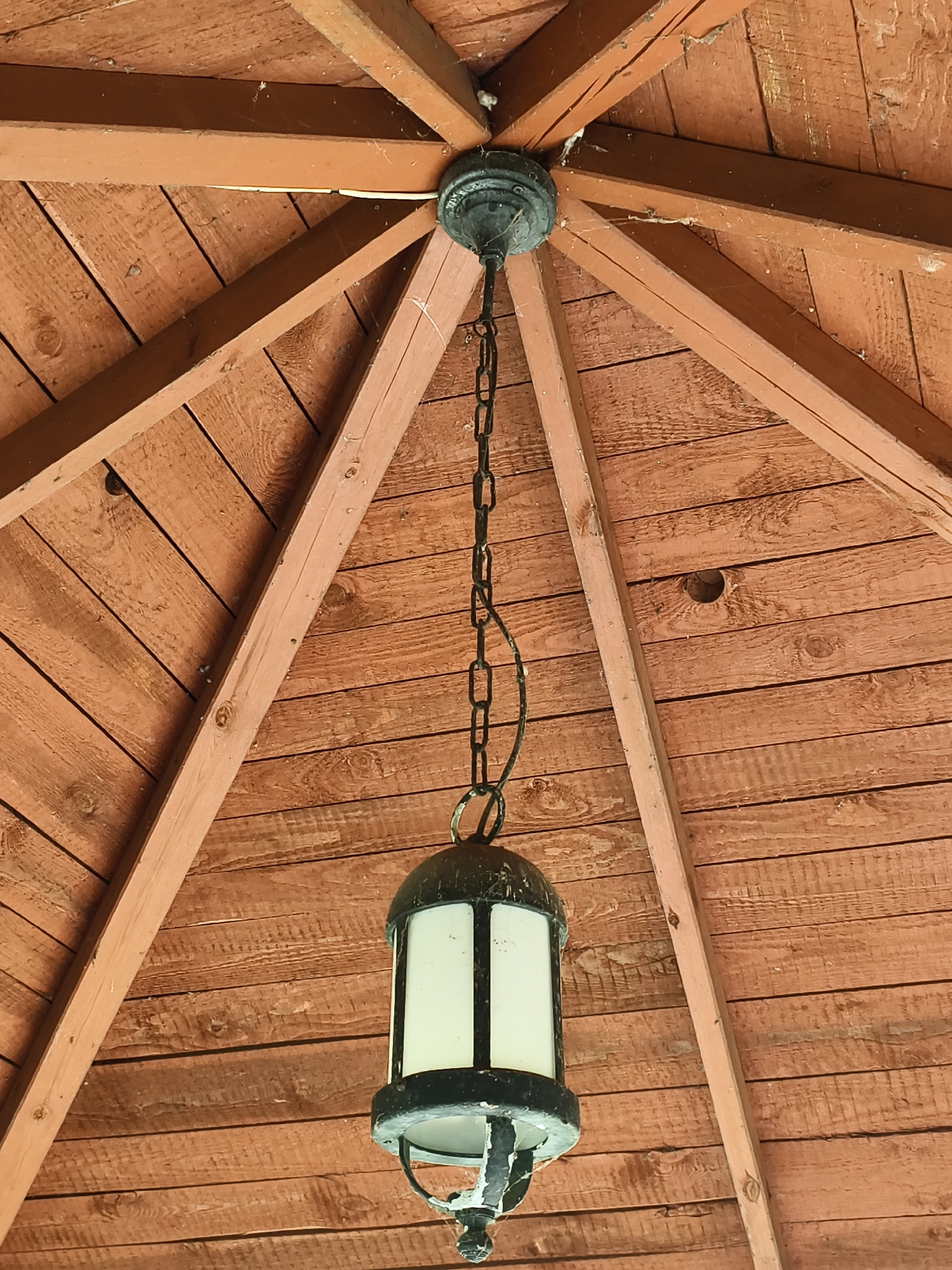 The image shows a close-up view of the wooden ceiling of an outdoor structure, possibly a gazebo or porch, with a hanging lantern. The ceiling is constructed of reddish-brown wooden planks laid horizontally, with triangular wooden beams radiating from a central point where the lantern is suspended. The lantern is a dark metal fixture with frosted glass panels. There are cobwebs visible in the corners and along the beams. The scene appears to be located in Tczew, Poland, based on the context provided. There are no people or animals visible in the image.