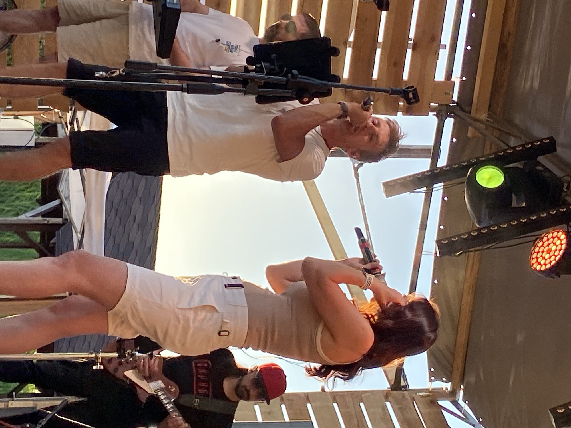 The image shows a performance taking place on an outdoor stage. In the foreground, a woman with long, dark hair is singing into a microphone, wearing a tan tank top and white shorts. Her left arm is raised, and she is looking upwards. To her left, a man wearing a black t-shirt, a red baseball cap, and a beard is playing a guitar.
Behind the woman, a man in a white t-shirt and black shorts is standing and singing into a microphone. He appears to be looking down at the woman. Above him, a tablet is mounted on a stand. The stage structure is made of wooden planks, suggesting an outdoor, possibly rustic or festival-like setting.
Lighting equipment is visible, including a green spotlight and a fixture with multiple red lights. The sun appears to be shining brightly, casting a warm light on the scene, and the sky is visible in the background, indicating it is daytime. The overall mood seems energetic, with the performers engaged in their music. There is no visible text in the image.