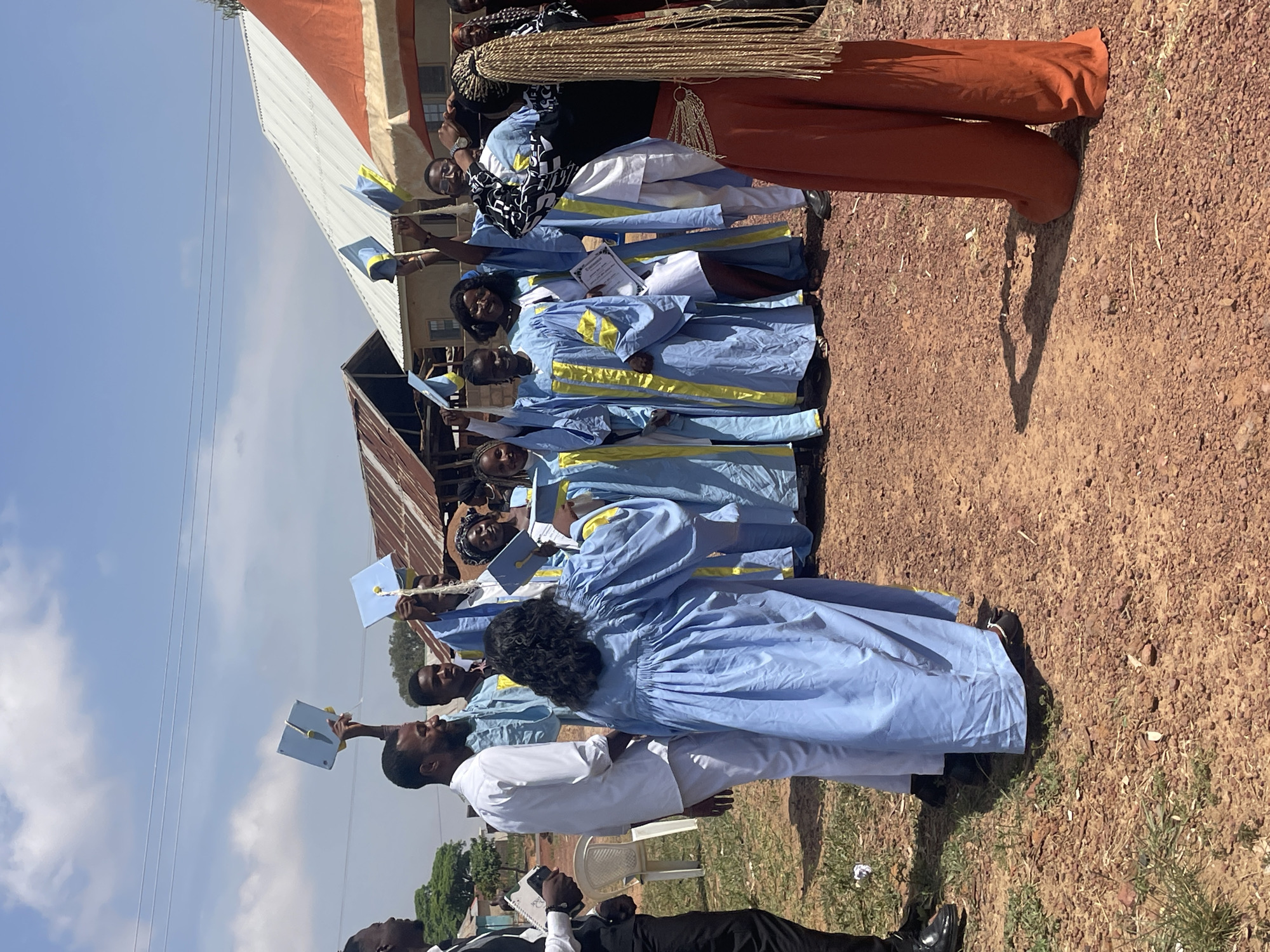 A group of people are standing outdoors in a rural setting, likely in or near Durumi, Nigeria, given the prompt information. The ground appears to be a mix of dirt and sparse vegetation. A building is visible in the background; its roof is constructed of corrugated metal. The sky is partly cloudy.
The main focus is on a group of people who seem to be celebrating an event. They are wearing light blue gowns with yellow accents and holding up objects, possibly diplomas or certificates. Some are also holding up blue graduation caps. Their facial expressions suggest they are happy and excited. Several people, including at least one wearing a white garment, are also present in the lower portion of the frame. One person in the front is wearing black pants and a black suit jacket.