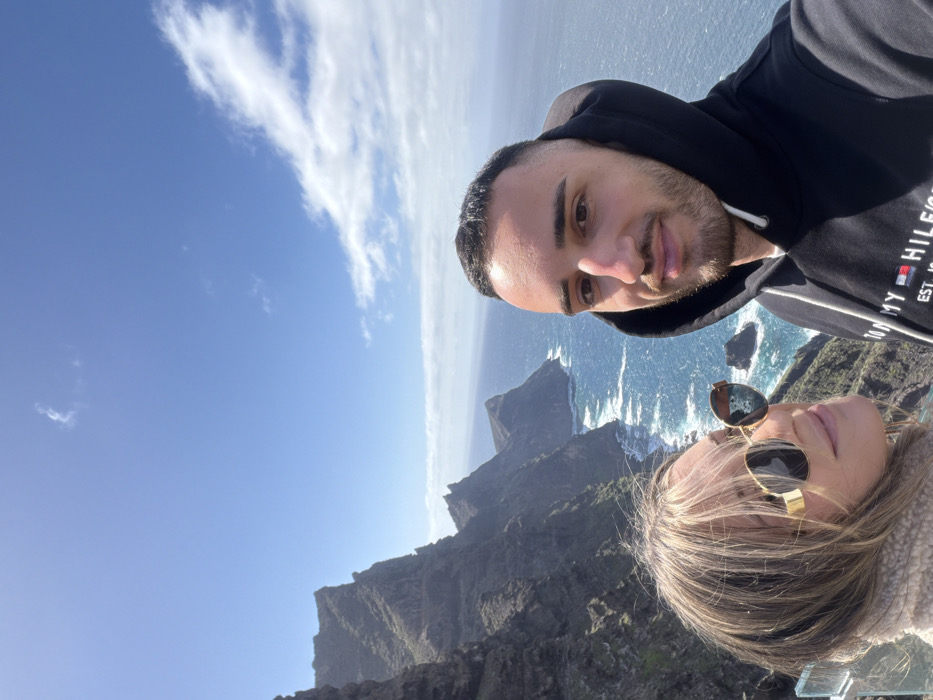 This image captures a selfie of two individuals, a male and a female, positioned at the Mirador del Balcón (also known as Mirador de Andén Verde) in La Aldea de San Nicolás, Gran Canaria, Spain. The male, on the right side of the frame, has short dark hair and a trimmed beard, wearing a black hooded sweatshirt with "TOMMY HILFIGER" visible on the chest. He looks directly at the camera with a slight smile. The female, partially visible on the left, has long blonde hair and is wearing round, gold-rimmed sunglasses and a light-colored, possibly beige, fuzzy top. She also looks towards the camera with a soft smile.
Behind them, the dramatic coastal landscape features rugged, dark volcanic cliffs that drop steeply into the deep blue Atlantic Ocean. White breaking waves are visible along the base of the cliffs and around smaller rock formations in the water. The sky above is clear and vibrant blue with a few scattered white clouds. A transparent glass barrier, indicative of a viewing platform, is partially visible in the lower left corner of the frame. The scene is brightly lit by natural sunlight.