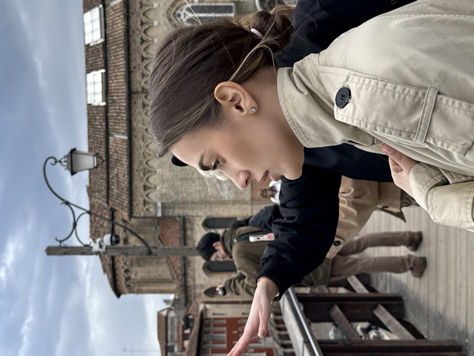 The scene depicts a group of individuals on a wooden bridge in Venice, Italy, specifically facing the Basilica dei Santi Giovanni e Paolo (San Zanipolo). The sky is overcast.
In the foreground, a young adult female is seen in profile, facing right. She has light brown hair tied back with a pink elastic band and wears a single circular stud earring. She is dressed in a light beige trench coat with a high collar and a visible black button on the right shoulder epaulet. Her right hand is near her chest, while her left arm is extended forward, palm open and fingers slightly spread, suggesting a gesture or pointing. Her mouth is slightly open.
Behind her, another individual's arm, dressed in black, is also extended forward, mirroring the gesture. Further back and to the left, a male figure in a dark green jacket and light brown trousers is holding a camera to his eye, apparently taking a photograph or video. A light-colored smartphone is attached to his back. Blurred figures of other individuals are present in the deeper midground.
The immediate environment includes wooden railings and decking of the bridge. A wooden pole with a decorative black lamppost and a white dome-shaped CCTV camera is positioned to the left of the background building. The prominent structure in the background is the Basilica dei Santi Giovanni e Paolo, characterized by its light stone and brick construction, arched windows, and tiled roof. To the far left, other multi-story Venetian buildings with tiled roofs and varied facades are visible along a canal.