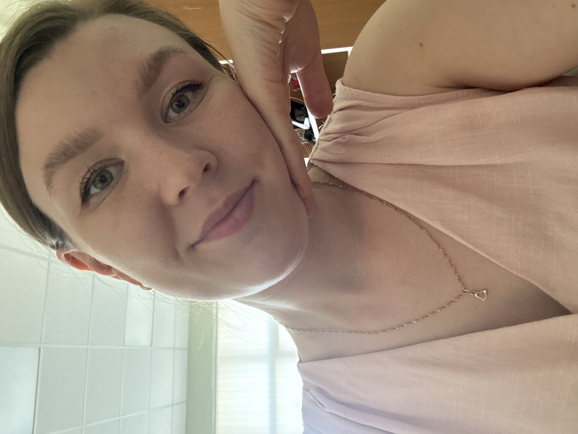 A selfie of a fair-skinned woman wearing a light pink top and a delicate gold necklace with a heart pendant. Her hand is near her face, partially obscuring her cheek. The background includes white tiled walls and a window, suggesting a bathroom setting. The location is not discernible from the image.