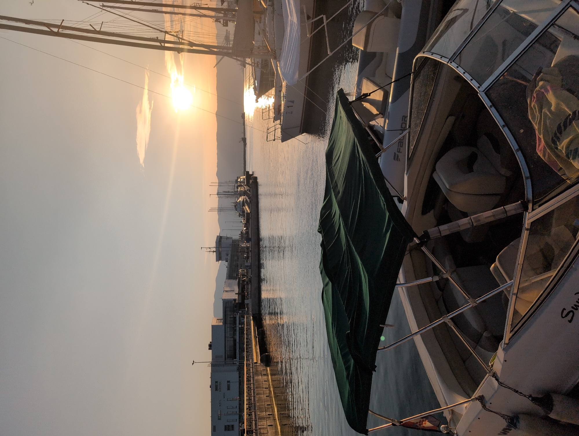 A marina in Rijeka, Croatia, at sunset. The sun is low on the horizon, casting a warm glow over the scene. Several boats are docked, including a small motorboat in the foreground with a green canopy. In the background, larger sailboats and other vessels are visible, along with industrial buildings and distant mountains. The water is calm and reflects the colors of the sky.