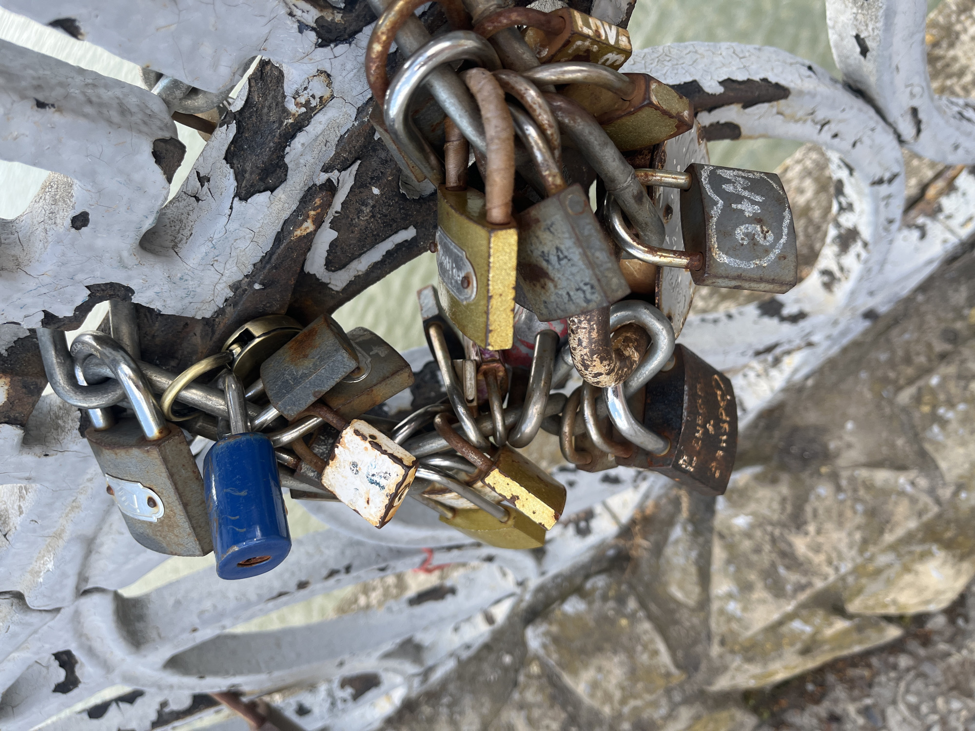 The image shows a close-up, overhead view of numerous padlocks attached to a white, weathered railing. The padlocks are of various shapes, sizes, and colors, with many showing signs of rust and wear. Some are brass-colored, others are silver or grey, and there is one prominent blue padlock. Many of the locks have chains looped through them, connecting them to the railing and to each other. The railing itself is a metal structure, painted white, but the paint is chipped and peeling, revealing darker metal underneath, suggesting age and exposure to the elements. The background is blurred, but appears to be a textured surface, possibly stone or concrete, with hints of green foliage. The overall impression is one of a "love lock" display, commonly found on bridges or railings, symbolizing commitment or remembrance. The lighting suggests it is daytime, with diffused light, possibly indicating an overcast day. No people are visible in the frame. Some locks have faint markings or inscriptions, but they are too indistinct to be clearly read in the image.