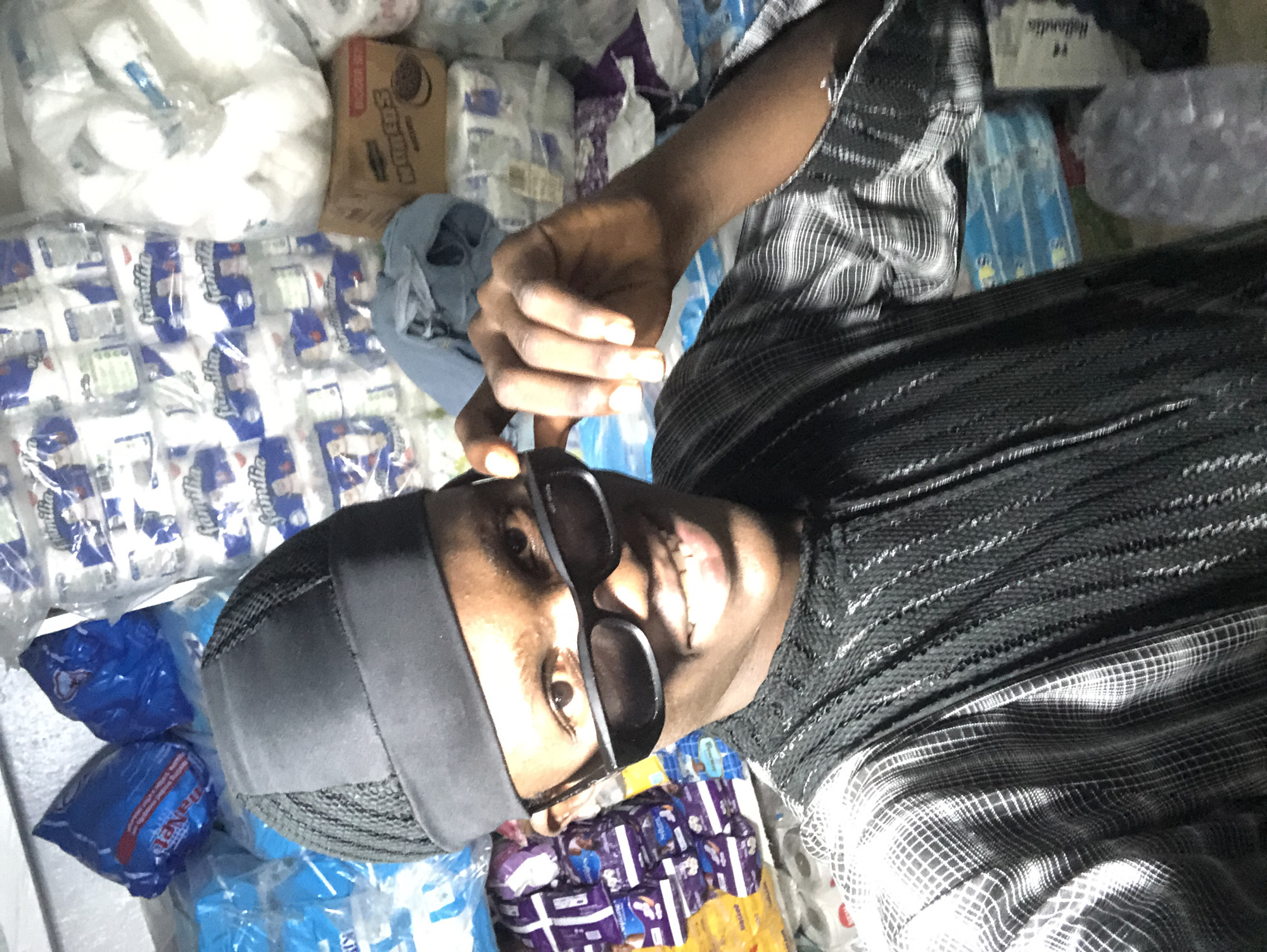 In Ado, Nigeria, a young Black male is pictured. He is wearing a dark kufi cap and dark sunglasses, and is dressed in a black and grey checkered outfit. He is smiling and holding his sunglasses up with his right hand. The background of the image is filled with stacked products, including packages of toilet paper and other goods, suggesting a retail or storage environment.
