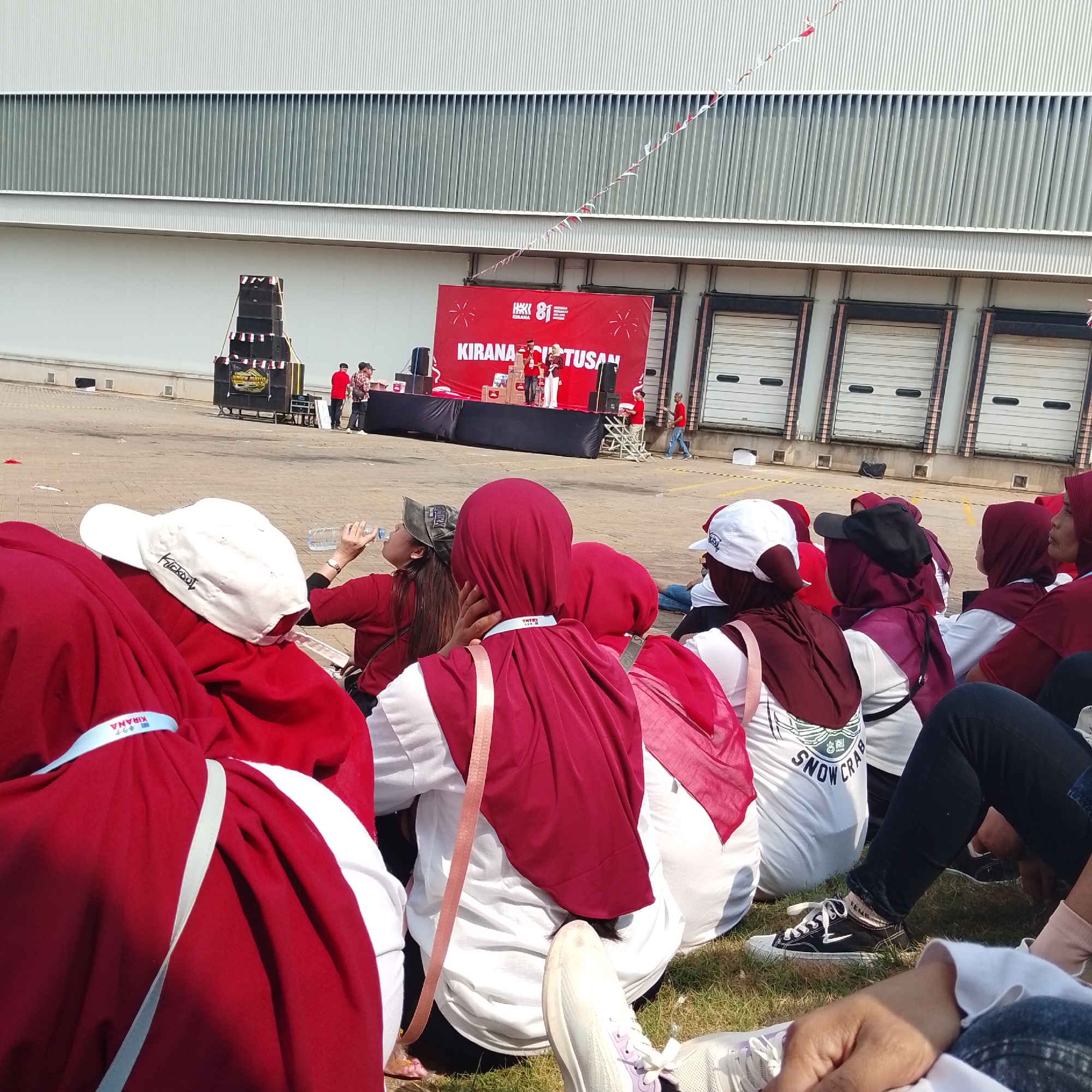 The image depicts an outdoor event in Plumpang, Indonesia, likely a company gathering or celebration. A group of people are seated on the grass in the foreground, facing a stage set up outdoors in front of a large, industrial-looking building with roll-up doors. The stage has a red backdrop with text that appears to be "KIRANA 81" and other Indonesian words. Two individuals are on the stage, one holding a microphone and another, possibly presenting an award or product. Several speakers and boxes are visible on and around the stage. The attendees in the foreground are mostly wearing red and white attire, with some wearing headscarves. The environment is sunny, with paving stones visible behind the seated crowd and the industrial building in the background. Several people are holding bottles of water. The overall atmosphere appears to be that of a casual, celebratory event.