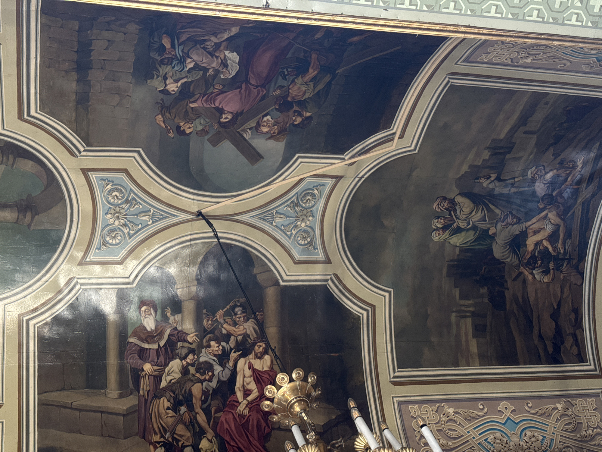 The image displays a fresco painted on a ceiling, depicting scenes from a religious narrative. The artwork is divided into several panels framed by ornate borders.
In the upper left panel, several figures in richly colored robes are gathered around a reclining figure. The reclining figure appears to be male, with his body partially draped in red cloth, and he has a beard and visible wounds. Other figures are leaning over him, some with expressions of distress or contemplation. The background in this section suggests an architectural setting with stone columns.
The upper right panel features a group of individuals struggling to carry a large wooden cross. A central male figure, wearing a purple robe, is being assisted by others who are pushing and pulling at the cross. The scene conveys a sense of effort and hardship. The background appears to be a dark, rugged landscape.
The lower panel showcases a group of figures in a mountainous or rocky terrain. Some are gathered around a fallen individual, who is lying on the ground. Other figures stand nearby, observing the scene. The background depicts a town or city with buildings silhouetted against a dimly lit sky.
Decorative elements, including geometric patterns and floral motifs, are visible in the framing of the panels. A chandelier or lighting fixture with multiple candle-like appendages hangs in the lower left portion of the image, partially obscuring one of the painted scenes. The overall style suggests classical or baroque religious art. The location is not specified in the image.