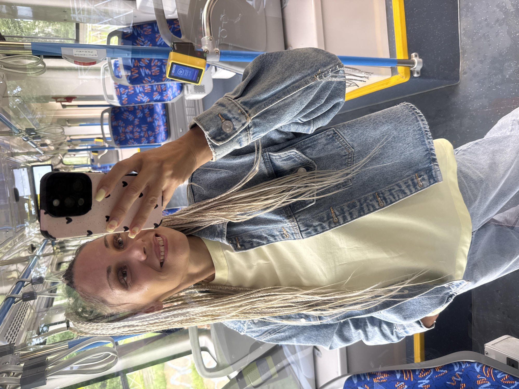 A woman with blonde braided hair is taking a selfie on what appears to be a public transport vehicle, possibly a bus or tram, as indicated by the blue seats with a patterned design and the presence of grab handles and poles. She is wearing a light yellow t-shirt and a denim jacket. Her phone, with a pink case adorned with black hearts, is visible in her hand. The interior of the vehicle suggests it is modern, with visible signage and a yellow handrail along the floor. The lighting suggests it is daytime. No specific location within Rusanovo, Russia, can be identified from the visual information.