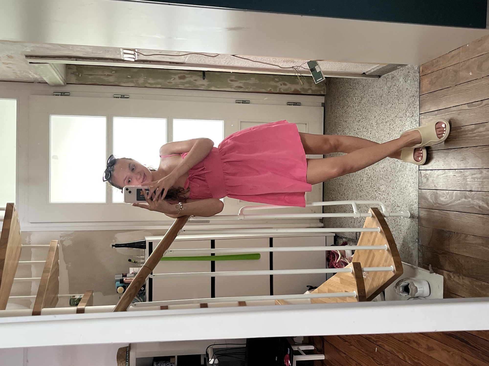 In this vertical, mirrored image, a young woman with dark hair pulled back and sunglasses perched on her head is taking a selfie in a hallway. She is wearing a bright pink, sleeveless dress and light-colored slide sandals. The woman is smiling at her phone, which she holds in her right hand. Her left arm is bent at the elbow, with her hand resting on the banister of a wooden staircase.
The setting appears to be a residential interior, likely in Saarbrücken, Germany. The hallway has a textured, light-colored wall on the left and wooden flooring on the right. A large set of white double doors with glass panes is visible in the background. The staircase, constructed of light-colored wood and white metal, is prominent in the foreground, reflecting in the mirror. Behind the staircase, a white dresser with multiple drawers is visible. There are some cleaning supplies and personal items on a shelf next to the dresser. The lighting suggests it is daytime. The overall mood is casual and relaxed.
