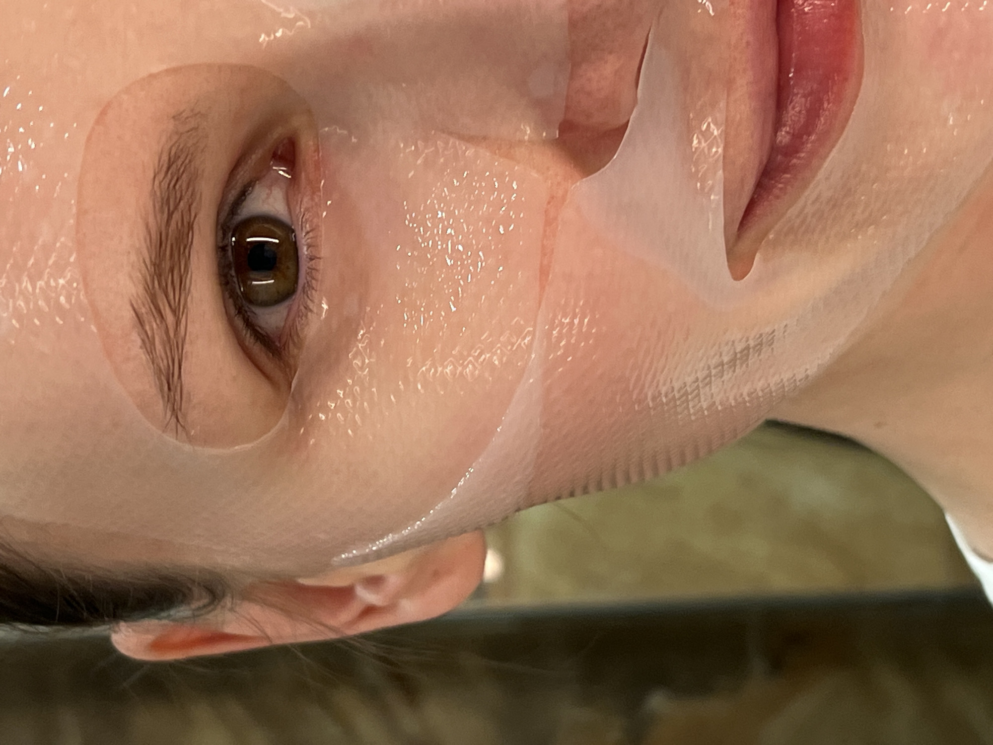 A close-up, rotated image shows a person's face with a sheet mask applied. The mask covers most of the face except for the eye, part of the ear, and the lips. The environment is blurry and out of focus. The location is Unknown, Unknown.