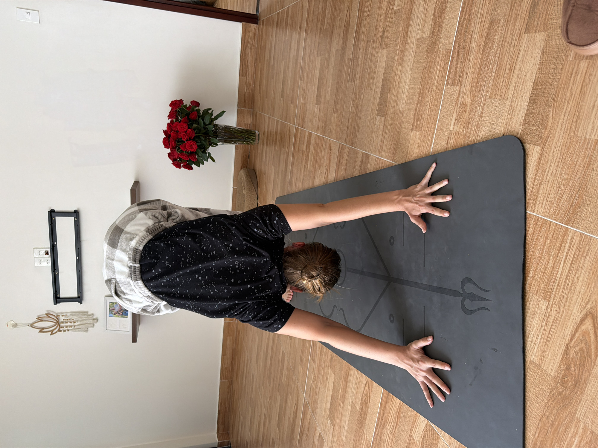 A person performs a yoga pose on a dark gray mat in what appears to be a room with light wood-tiled flooring. The person is wearing a black t-shirt with white star patterns and a plaid shirt draped over their shoulders. Their head is lowered, and their hands are flat on the mat, with fingers spread. The background shows a white wall with a framed picture and decorative items. A vase of red roses sits on a surface to the right. The image does not contain any specific geographical indicators that would allow for the identification of the location as Lâm Viên - Đà Lạt, Vietnam.