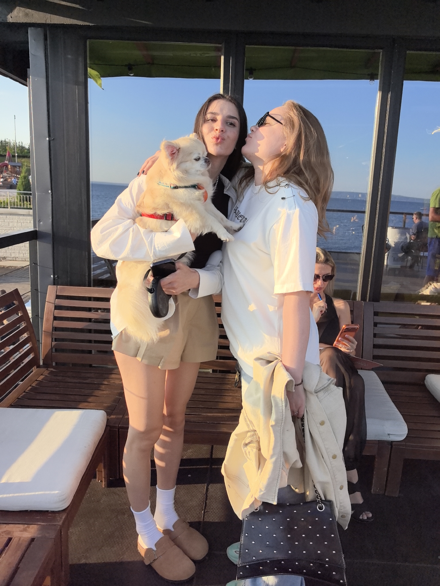 Two young women are holding a small, light-colored dog on a covered outdoor patio with a view of the Volga River. The woman on the left is wearing a white shirt and tan shorts, holding the dog in her arms and puckering her lips. The woman on the right is wearing a white t-shirt and sunglasses, leaning in to kiss the dog. The dog is wearing a red collar and a leash is attached. Behind them, a woman in sunglasses is sitting on a wooden bench and looking at her phone. The patio appears to be part of a restaurant or cafe on the embankment of the Volga River in Tolyatti, Russia. The sun is setting, casting a warm glow on the scene.