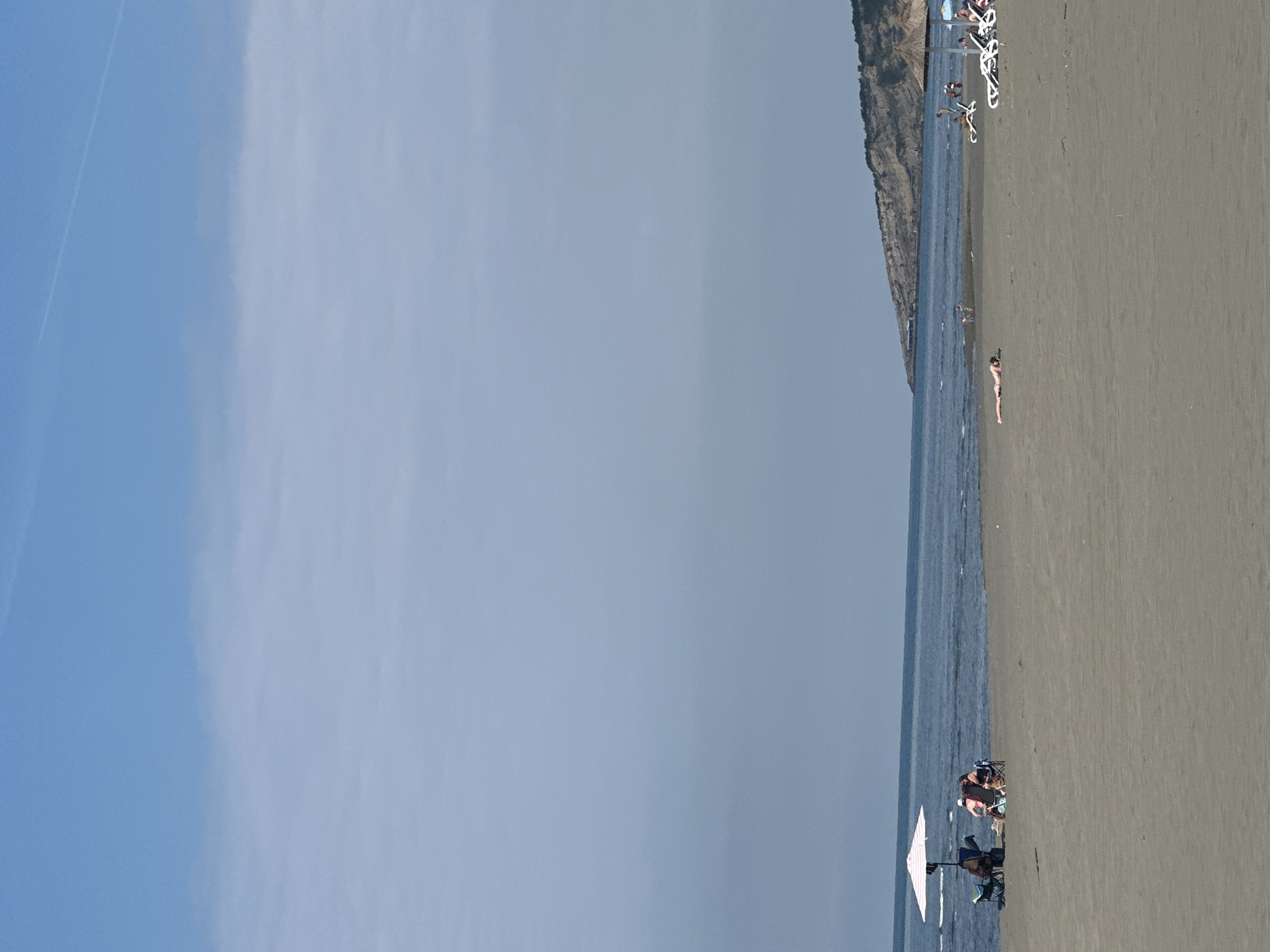The scene is a beach in Donji Štoj, Montenegro. The image captures a wide sandy beach, a strip of blue water, and a sky that transitions from a bright blue to a hazy grey. Along the shoreline, a cliff is visible. People are sparsely spread along the beach. Some are relaxing under an umbrella, and one person is lying down near the water. Beach chairs and umbrellas are scattered along the beach.