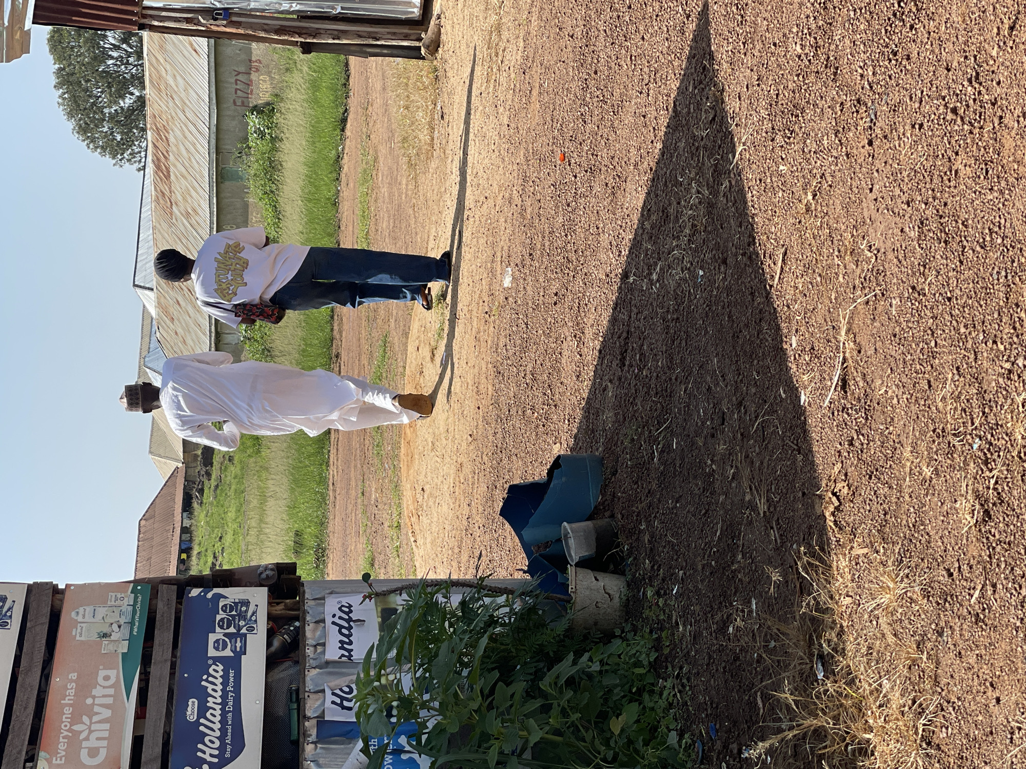 This media file captures a scene in Durumi, Nigeria, under a clear, sunny sky, likely in the late morning or early afternoon given the long shadow cast by a person. Two individuals are visible. The foreground shows a person from the waist down, dressed in a traditional white garment and a cap, walking away from the camera. Behind them, another person is also walking away, wearing a white sweatshirt with yellow lettering on the back and blue jeans. They are carrying a bag with a red and black patterned strap.
The setting appears to be an outdoor area with a rough, reddish-brown gravel ground. In the background, there are corrugated metal buildings and green vegetation, suggesting a semi-rural or mixed-use area. Several signs are displayed on wooden planks. One sign reads "Everyone has a Chivita" and another features the "Hollandia" brand with the tagline "Stay Ahead with Dairy Power". Other partially visible text includes "FIZZY", "IRISCO", and "Ho India". There are also some scattered plastic objects and a broken blue container on the ground. The overall impression is a casual outdoor environment in a Nigerian locality, with commercial signage adding to the visual context.
