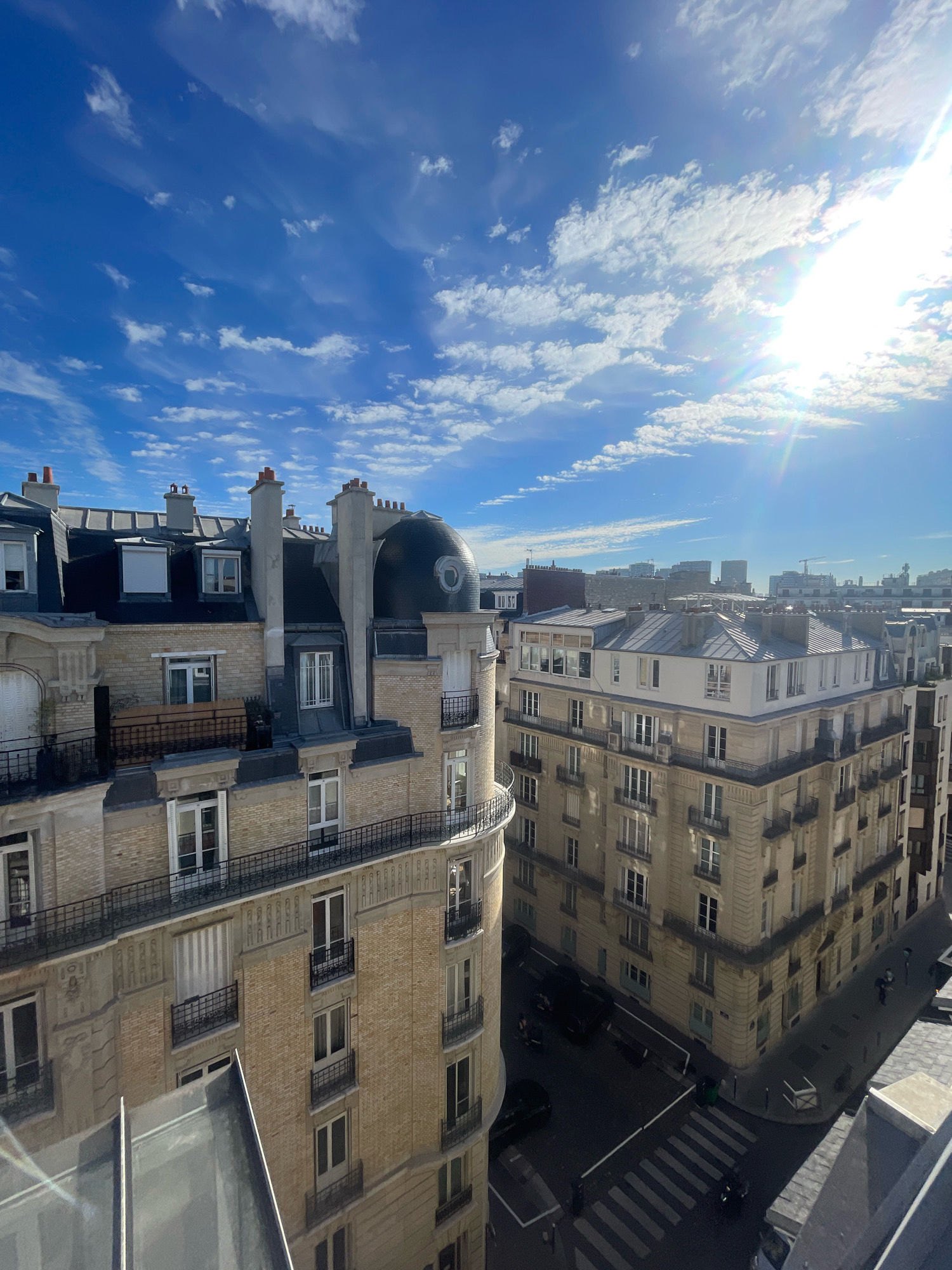 The image is an exterior, elevated view of Parisian apartment buildings on a sunny day. The sky is bright blue with scattered white clouds and the sun is shining intensely in the upper right quadrant, creating lens flare. In the foreground, a portion of a light-colored building with a slanted roof is visible. To the left and center, a multi-story building with a light beige brick facade, dark mansard roof, and dormer windows is prominent. It features balconies with black railings on several floors, and ornate decorative elements above some windows. To the right, a larger, taller building with a similar beige facade and multiple rows of windows is visible. This building has a mansard roof and is situated on a street corner. The street below is paved, with a pedestrian crossing and several dark-colored cars parked or driving. Some people are visible walking on the sidewalk. In the background, more buildings of varying heights can be seen, characteristic of a dense urban environment. The architecture suggests the image was taken in Paris, France, likely in an older, established neighborhood.