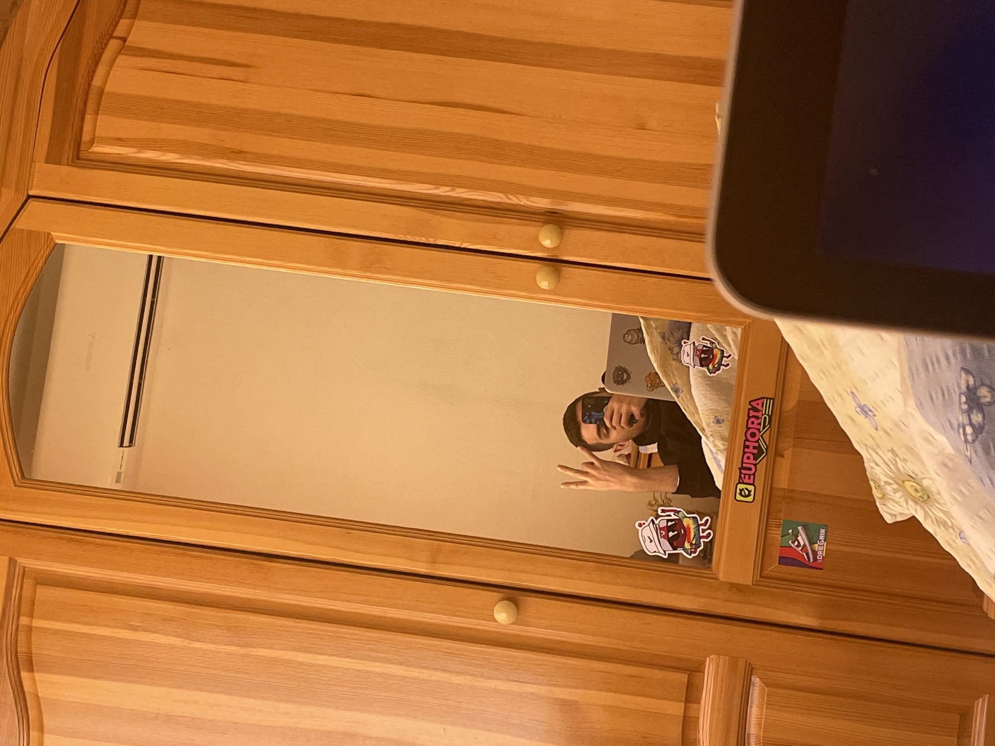 A selfie is being taken by a person reflected in a wardrobe mirror. The wardrobe is wooden with decorative panels and round knobs. The person in the reflection is holding a phone to take the photo, and their face is visible. The background includes a bed with patterned bedding and a white air conditioning unit mounted on the wall. Stickers are attached to the side of the wardrobe. The location may be Elena, Bulgaria.