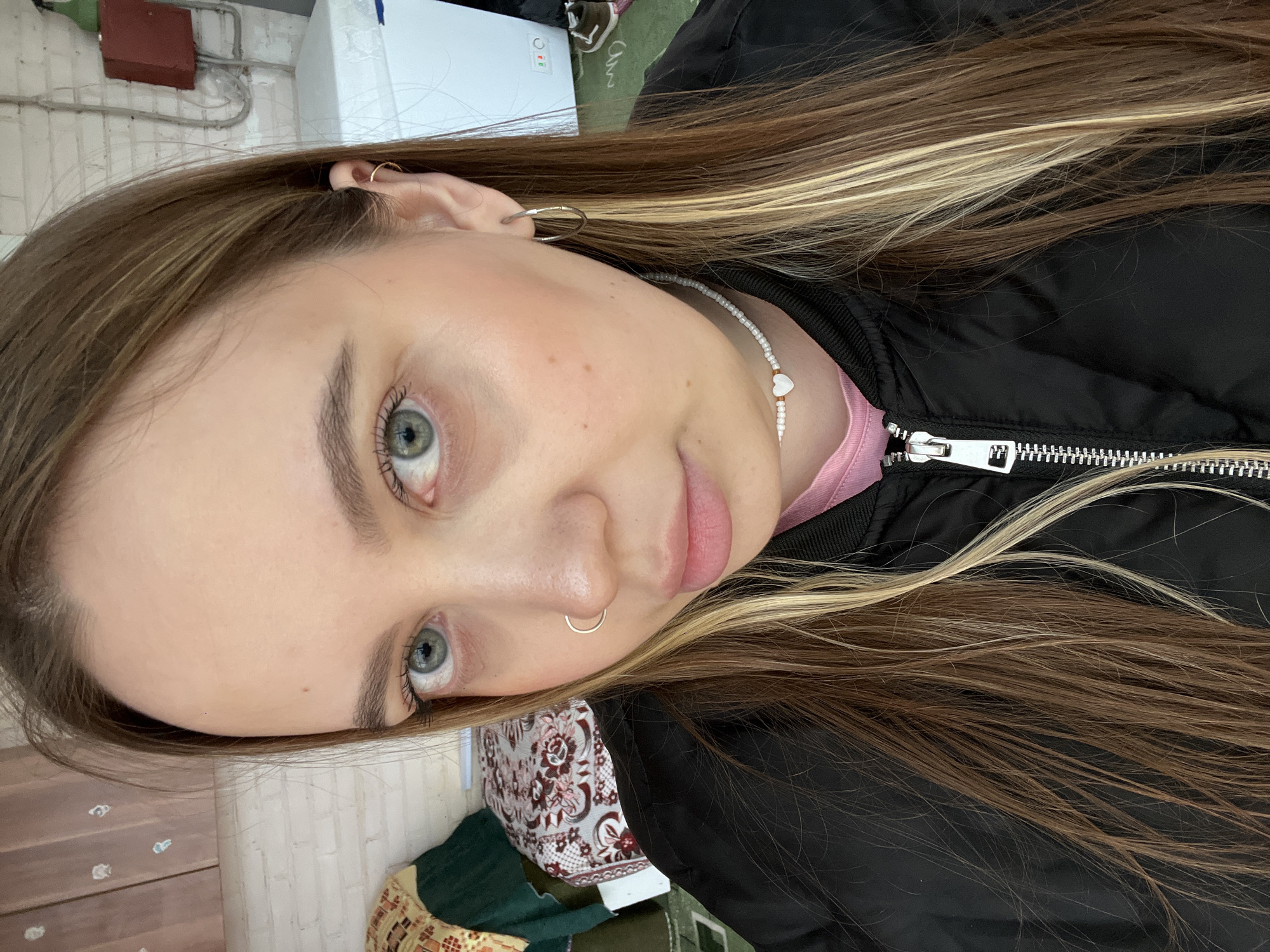 A selfie taken indoors, likely in Novopokrovskaya, Russia. The subject is a young woman with long, brown hair streaked with blonde highlights. She has light skin, light-colored eyes, and is wearing earrings and a nose ring. She is wearing a black jacket over a pink shirt and a beaded necklace with a heart charm. The background includes a brick wall, pipes, a white appliance, and miscellaneous items on a surface. The photo is rotated.