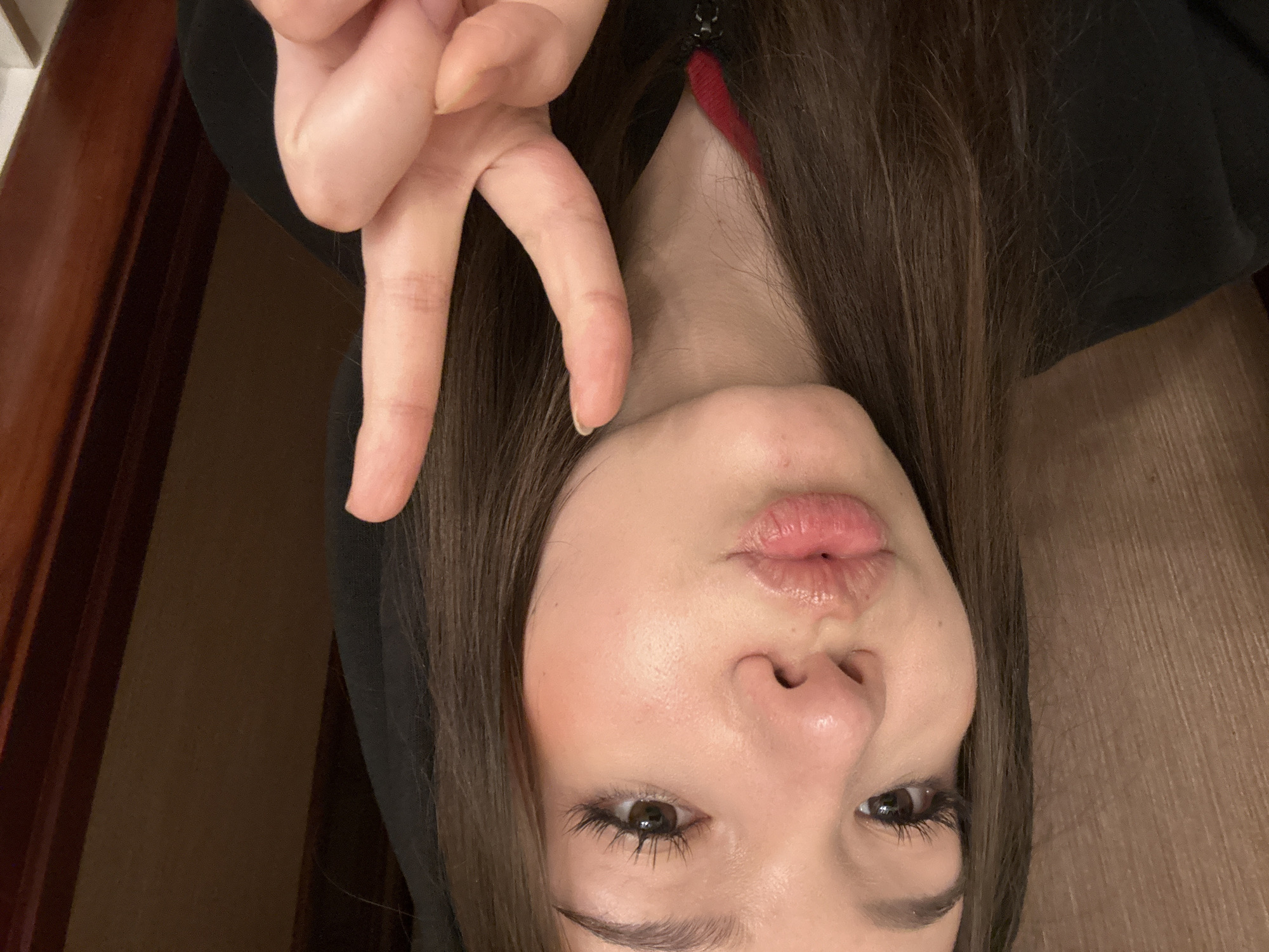 A young female is depicted in a close-up indoor selfie. She has long, dark brown hair and is wearing a black hooded sweatshirt with a visible red accent near the neckline zipper. Her face is directed towards the camera, with her lips puckered in a kissing pose. Her right hand is raised, forming a peace sign with the index and middle fingers extended. Her eyes are dark brown, featuring prominent eyelashes. The background on the left consists of a light brown, vertically textured wall or paneling, while the right side shows a darker brown wall and a visible wooden doorframe. The specific location within the city of Naberezhnye Chelny, Russia, cannot be determined from the visual information present in this image.