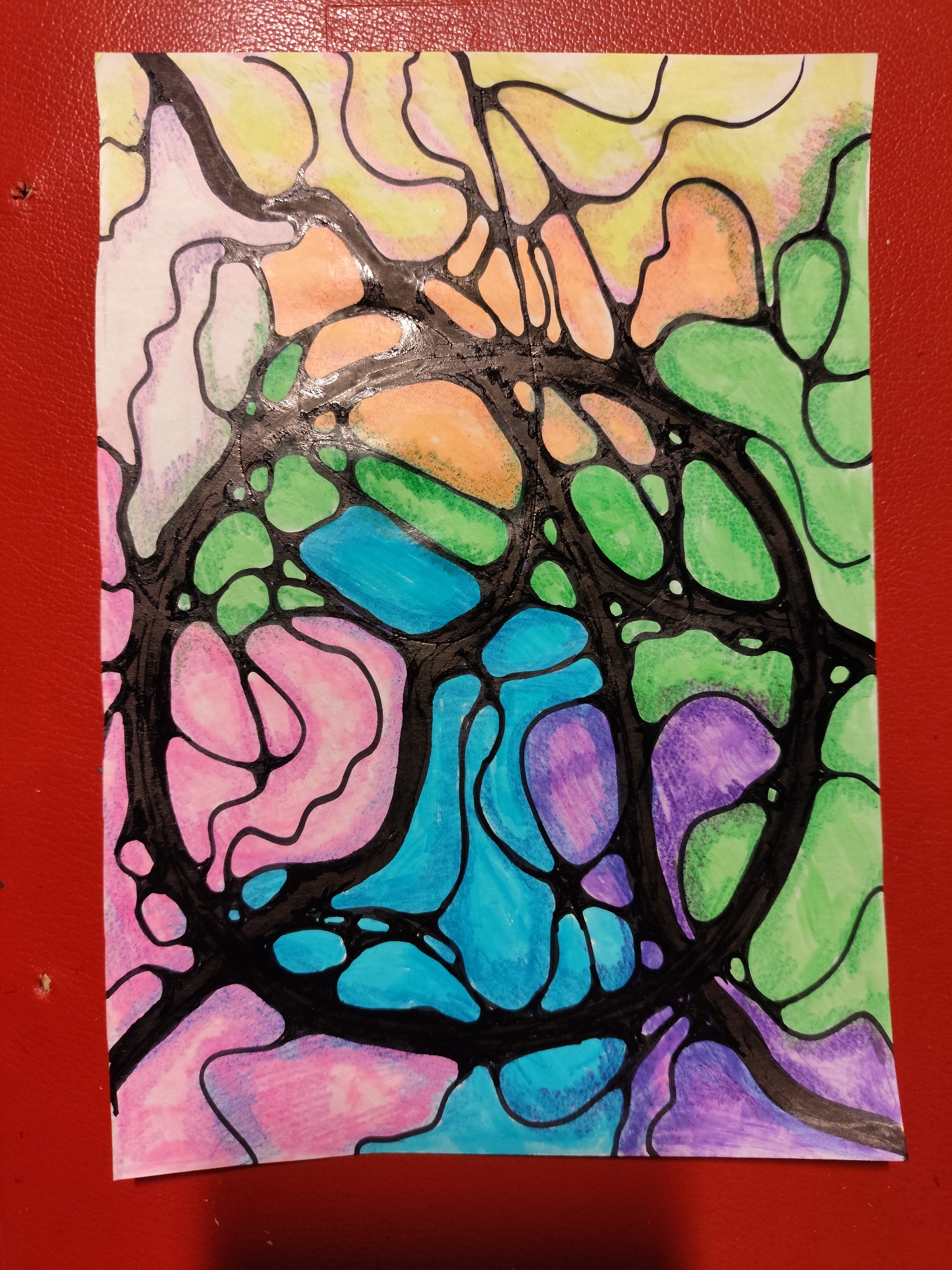 The image shows a piece of paper with abstract artwork, possibly a neurographic drawing. The artwork features interconnected black lines forming organic shapes that are filled with various colors including pink, purple, blue, green, orange, and yellow. The paper is placed on a textured red surface, likely a table or a mat. There is no indication of the city of Krasnodar, Russia, or any identifiable landmarks within the image.
