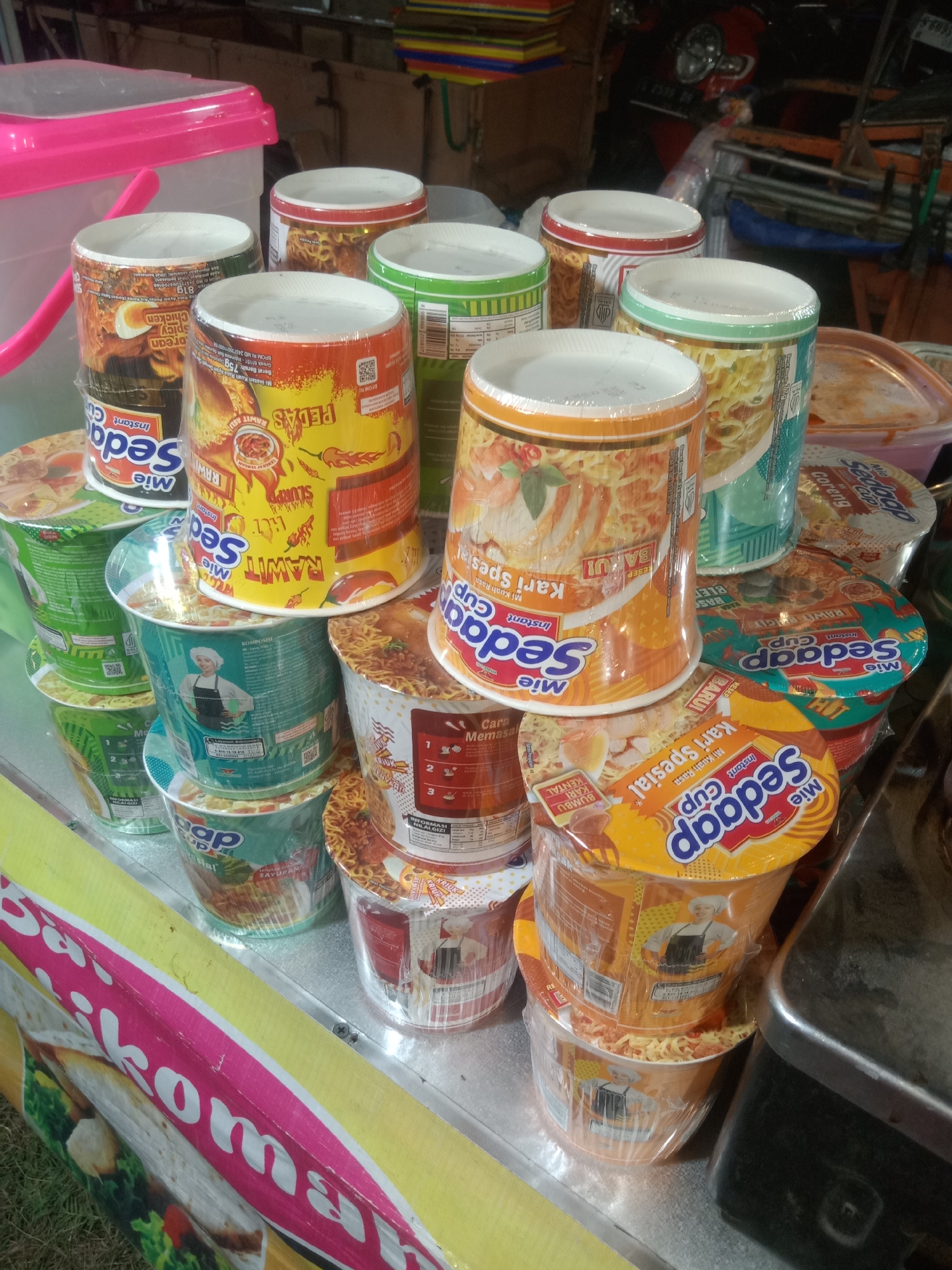 In Stiaji, Indonesia, a close-up shot captures an outdoor market stall displaying a variety of instant noodle cups. The noodles are stacked on a metallic surface, with a bright yellow and pink banner, possibly advertising "Balikoman", visible in the foreground. The stall is dimly lit, with various food items and containers visible in the background, including a pink plastic container and a red motorcycle. The noodle cups, primarily branded "Mie Sedaap", are individually wrapped in clear plastic. Different flavors and varieties are visible, indicated by the colorful packaging featuring images of food and text in Indonesian. The focus is on the detailed presentation of the noodle products, highlighting their different flavors and preparation instructions.