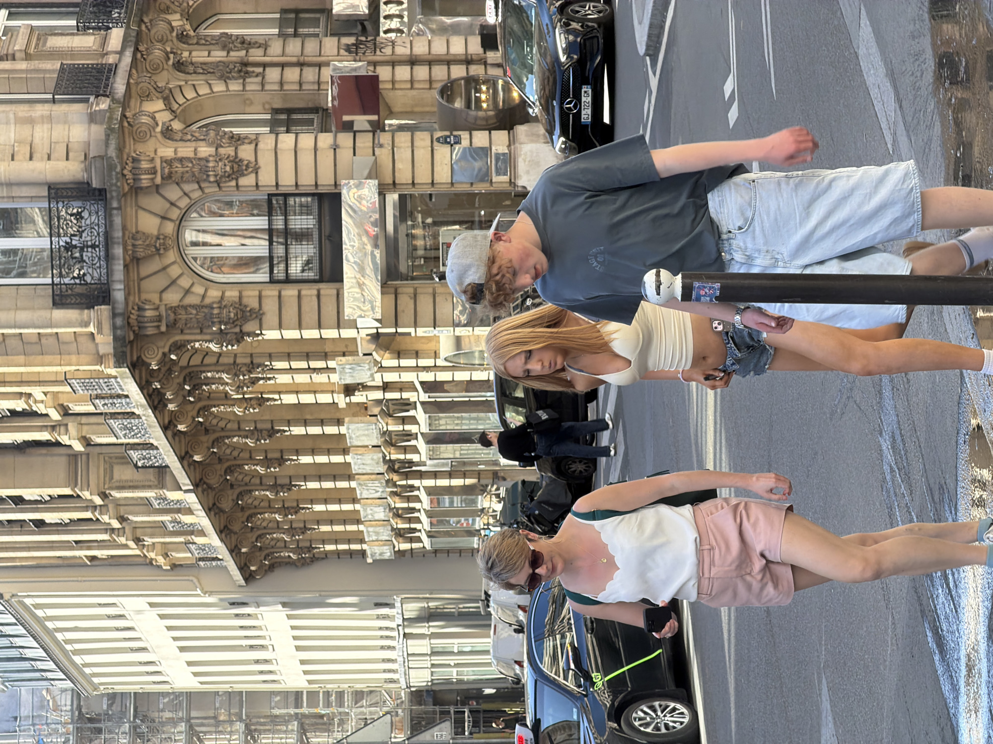 The photograph captures a street scene in Paris, France, evidenced by the distinctive architecture of the buildings in the background. Three individuals are prominent in the foreground. A young man wearing a gray t-shirt, light blue denim shorts, and a backward baseball cap stands beside a woman with blonde hair, a white top, and short denim shorts. In front of them is a woman with short hair, sunglasses, a white top with green straps, and pink shorts, holding a phone.
The environment includes parked cars, pedestrians in the background, street markings, and a metal street barricade on the right side of the frame. The overall scene is a well-lit, daytime street view in a European city.