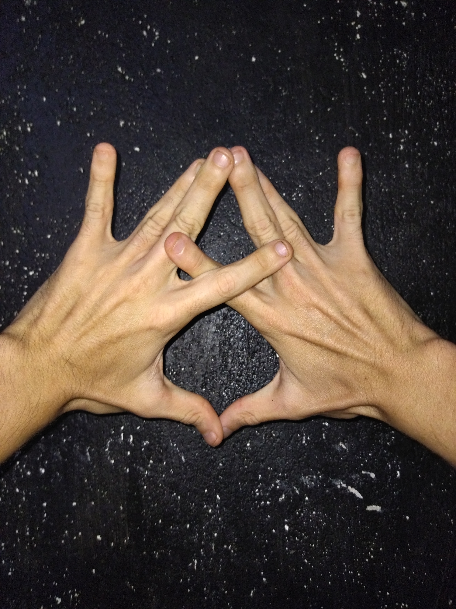 The image shows two human hands interlocked to form a triangular shape, against a dark, textured surface with small white speckles. The hands appear to be fair-skinned, with visible veins and hair. The fingers are extended, and the thumbs are crossed. No other people, subjects, objects, or actions are discernible in the image. There is no information present in the image that can identify the location as Tinaquillo, Venezuela, or any other specific place.