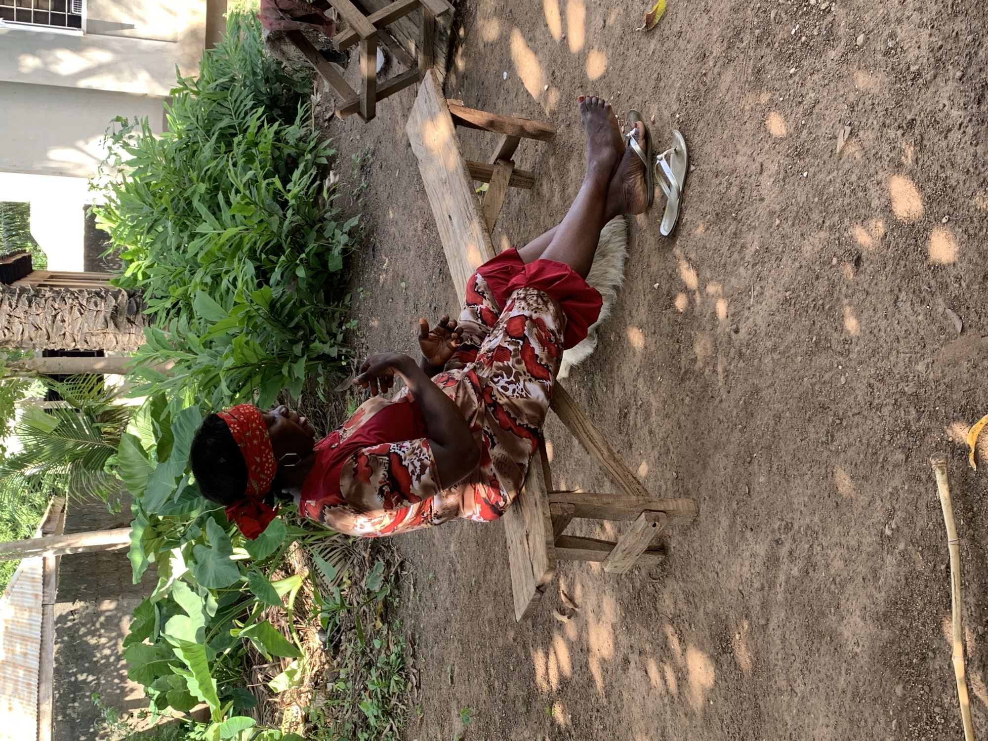 An African woman wearing a red and brown patterned dress and a red headscarf sits on a weathered wooden bench outdoors. Her feet, adorned with red nail polish, rest on flip-flops on the ground. She is in a shaded area with a rough, dirt ground and scattered leaves. In the background, lush green plants and trees are visible, along with a corrugated metal roof on a building. A wooden frame structure is partially visible to the right. The scene appears to be a rural or semi-rural setting within the city of Akwanga, Nigeria, likely a residential or garden area.