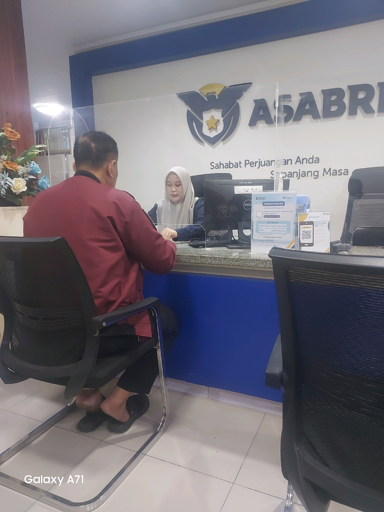 In a well-lit indoor environment, likely a service center or office, a male customer wearing a maroon jacket and black pants sits facing away from the camera in a black office chair. He is interacting with a female employee who is behind a counter, separated by a clear partition. The employee, wearing a light-colored hijab and a dark top, is seated and facing the customer. On the counter are computer monitors, a clear plastic stand with information, and other office supplies. Behind the counter, a large sign displays the ASABRI logo and slogan "Sahabat Perjuangan Anda Sepanjang Masa". The floor appears to be tiled. The overall scene depicts a typical customer service interaction.