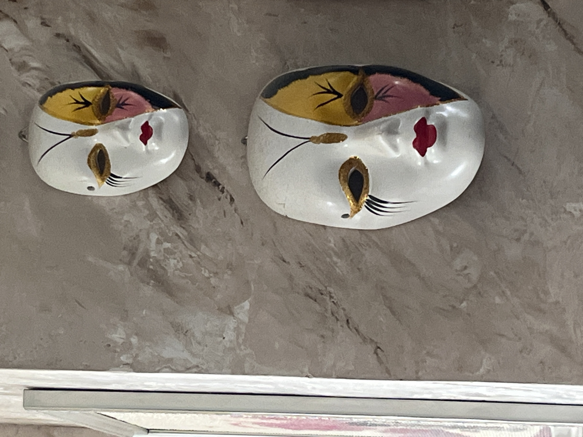 The image shows two decorative masks hanging on a textured wall. The masks are white with a half-face design in yellow, pink, and black, resembling butterfly wings. They have gold accents around the eyes and red lips. The wall has a marbled pattern in shades of beige and gray. To the left of the wall, a portion of a white frame is visible, suggesting it might be a picture frame or part of a doorway. The scene appears to be indoors, likely a residential setting. There is no information in the image that would allow for identifying the city of Vladimirovka, Russia.