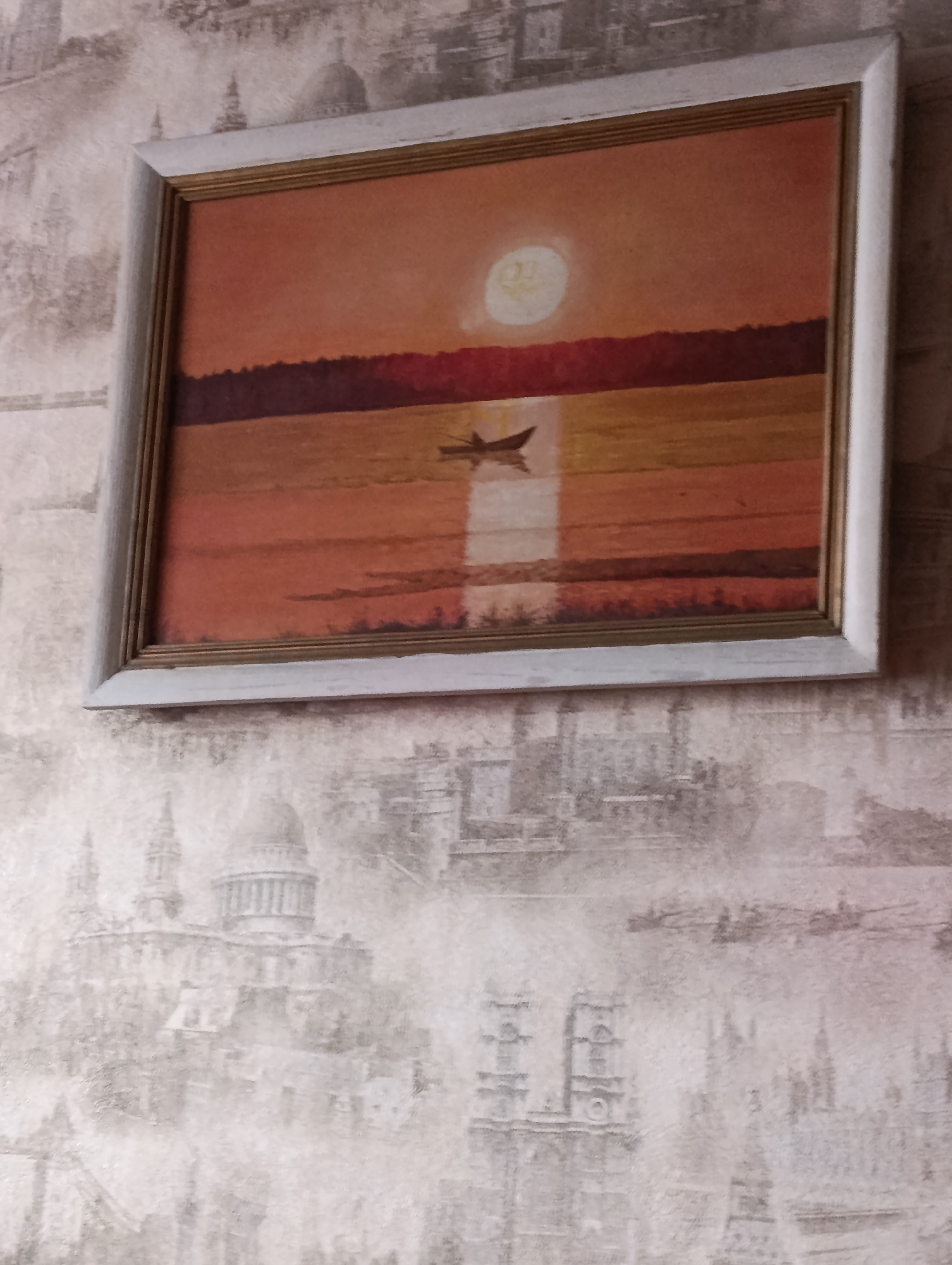 The image shows a framed painting hanging on a wall covered in patterned wallpaper featuring faint architectural scenes. The painting depicts a sunset scene with a body of water, a distant treeline, and a single boat. The sky is orange with a bright circular object, likely the sun, situated toward the upper center. The painting is framed in a light-colored frame with a thin inner gold border. There are no people visible. The location is not determinable from the image content alone.
