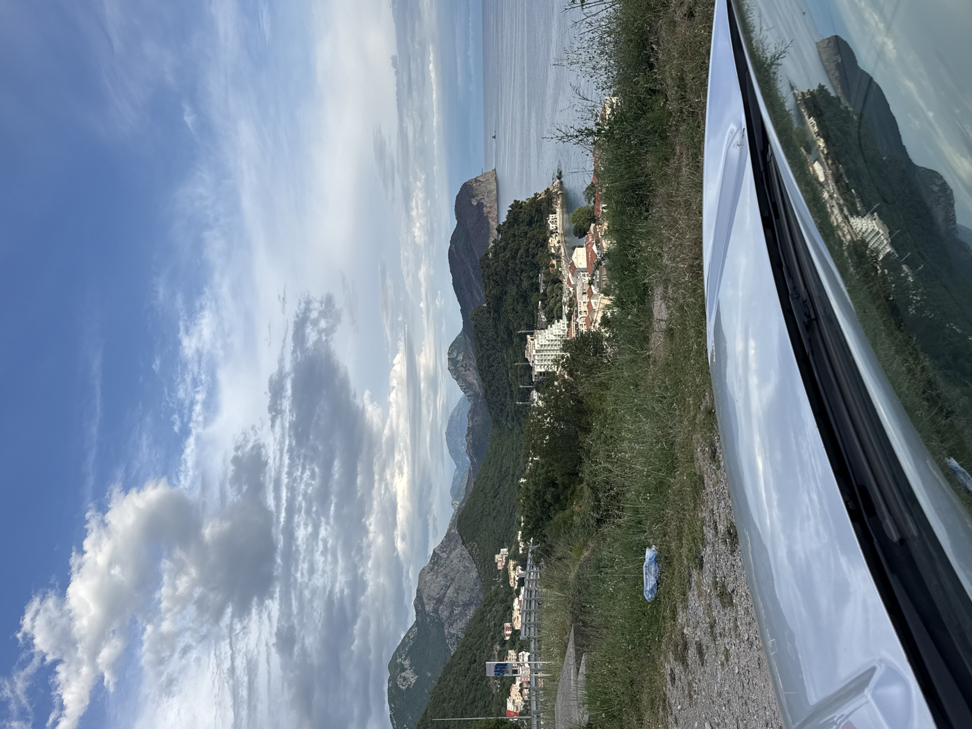 The image shows a view from a high vantage point overlooking the city of Petrovac na Moru, Montenegro. A car is partially visible on the right side of the frame, with the landscape reflected in its windows. In the distance, the Adriatic Sea stretches out to the horizon. In the foreground, the landscape slopes down towards the city, composed of buildings nestled among the hills and coastal area. Mountains rise in the background, creating a layered effect of hills and peaks. The sky is partly cloudy. Litter is visible on the side of the road.