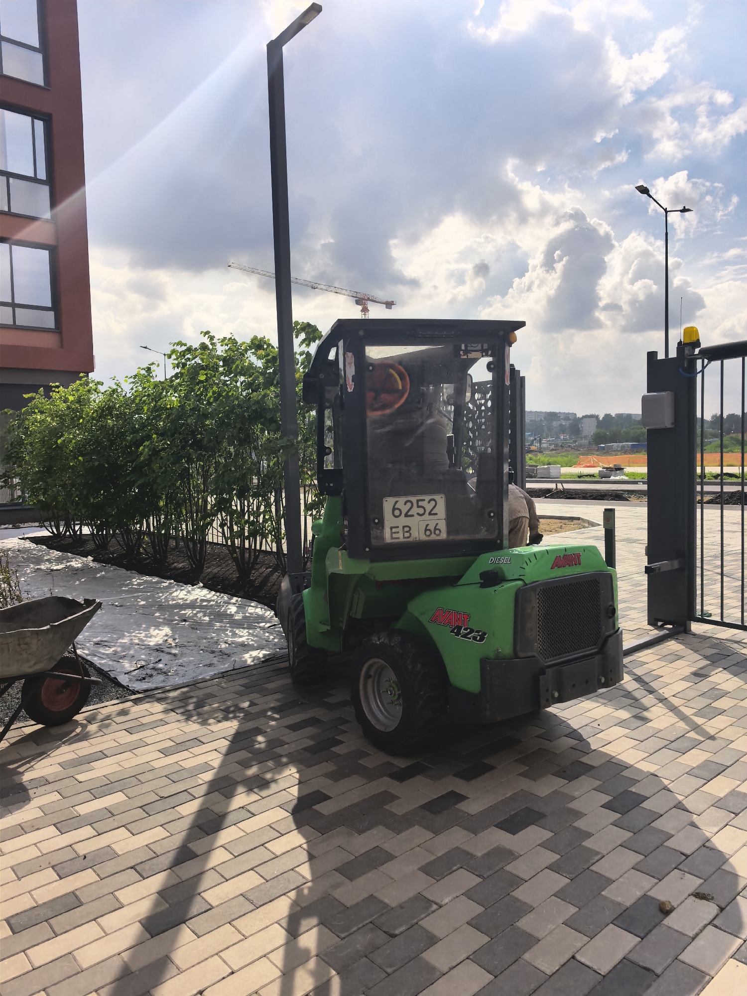 The image depicts an outdoor scene in the city of Unknown, Unknown, characterized by active landscaping or construction. In the immediate foreground, a multi-toned paved walkway, composed of rectangular pavers in shades of beige, light gray, and dark gray, extends across the lower portion of the frame. Long, distinct shadows, cast by objects off-frame and a central street light pole, stretch diagonally across the paving from the upper right.
On the left side of the foreground, a metal wheelbarrow with an orange wheel is partially visible. Adjacent to the paved area and the wheelbarrow, a dark ground cover material is visible, partially covered by a white sheet of what appears to be geotextile or mulch fabric. A dense row of green bushes or small trees is planted in dark soil behind this ground cover, extending across the middle ground.
Centrally located, a tall, black, slender street light pole stands vertically, casting a prominent shadow. To its right, a green Avant 423 compact multi-loader is positioned, facing slightly away from the viewer. The loader bears "DIESEL" and "AVANT" branding, and a rear license plate displaying "6252 EB 66". A person wearing a light-colored top is visible operating the loader from within its cabin. Another person is partially obscured behind the loader.
To the right of the loader, a black metal gate with vertical bars is partially open. It is equipped with an automated opener mechanism mounted on its post and a yellow flashing beacon light on top.
In the far left background, a portion of a modern building with reddish-brown exterior cladding and dark-framed windows is visible. The sky above is partly cloudy with significant sunlight, creating a lens flare in the upper left quadrant. In the distant background, indications of a larger construction site are visible, including at least two red and white tower cranes, various earthworks, and additional distant structures under construction. Another street light fixture is visible in the middle-distance background.