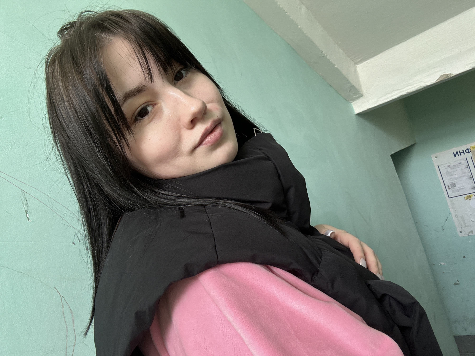 A young woman with black hair and bangs is posing for a selfie indoors. She is wearing a pink top and a black puffer jacket. The background is a mint green wall with a sign in Russian that reads "ИНФ" partially visible. The setting appears to be a hallway or stairwell in a building. The location is in the city of Chaikovsky, Russia.
