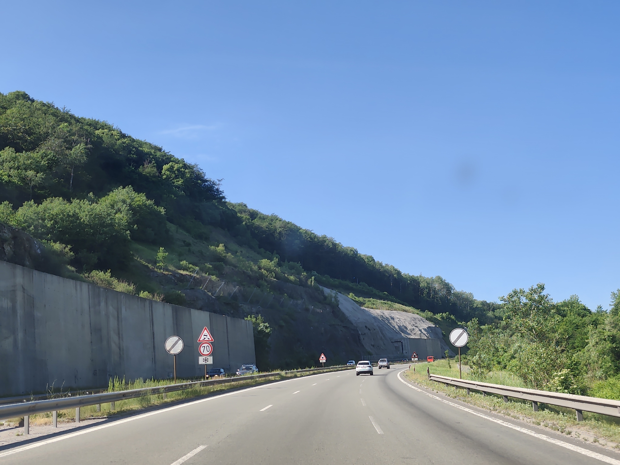 The image depicts a highway with multiple lanes traveling through a mountainous, tree-covered area. A concrete retaining wall runs along the left side of the road, supporting a steep, forested slope. Various traffic signs are visible along the roadside, including a speed limit sign indicating 70 and a warning sign for slippery road conditions. Several cars are driving on the highway. The sky is clear and blue. The location is identified as Malo Buchino, Bulgaria.