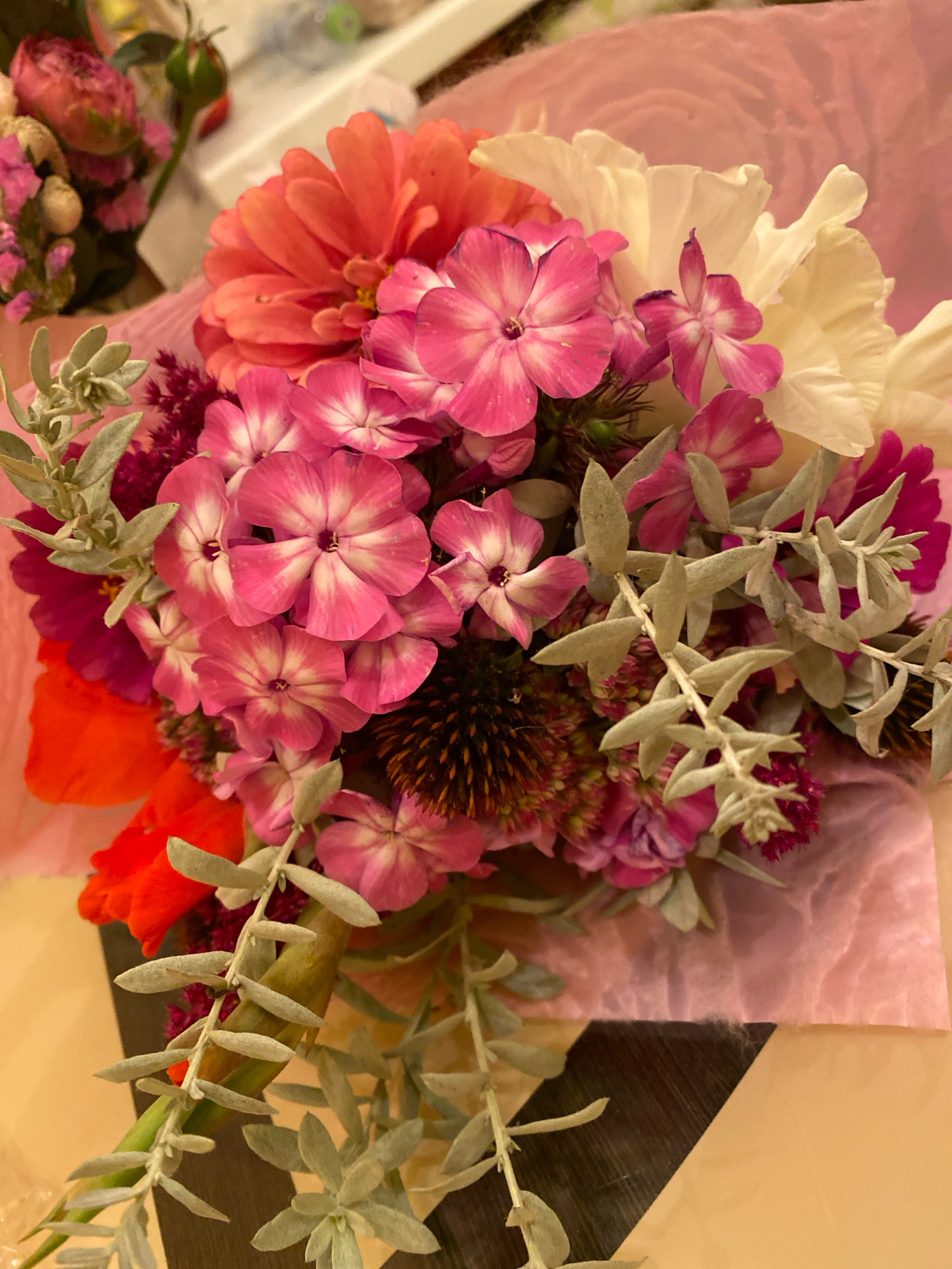 A close-up, overhead shot shows a bouquet of flowers wrapped in pale pink tissue paper. The bouquet is arranged with a variety of flowers in shades of pink, orange, and white, interspersed with silver-grey foliage. Prominently featured are bright pink, star-shaped flowers with white centers, a large coral-colored zinnia, and some delicate white, ruffled petals. Smaller clusters of dark pink and burgundy flowers add depth. The overall impression is one of fresh, vibrant floral arrangement. The background consists of a surface with a diagonal pattern of light brown and dark wood-like texture, suggesting a table or shelf. The lighting is bright, illuminating the flowers and bringing out their colors and textures. There are no people or animals visible in the image. This image does not contain any information that can be used to determine the location in Ivanteyevka, Russia.