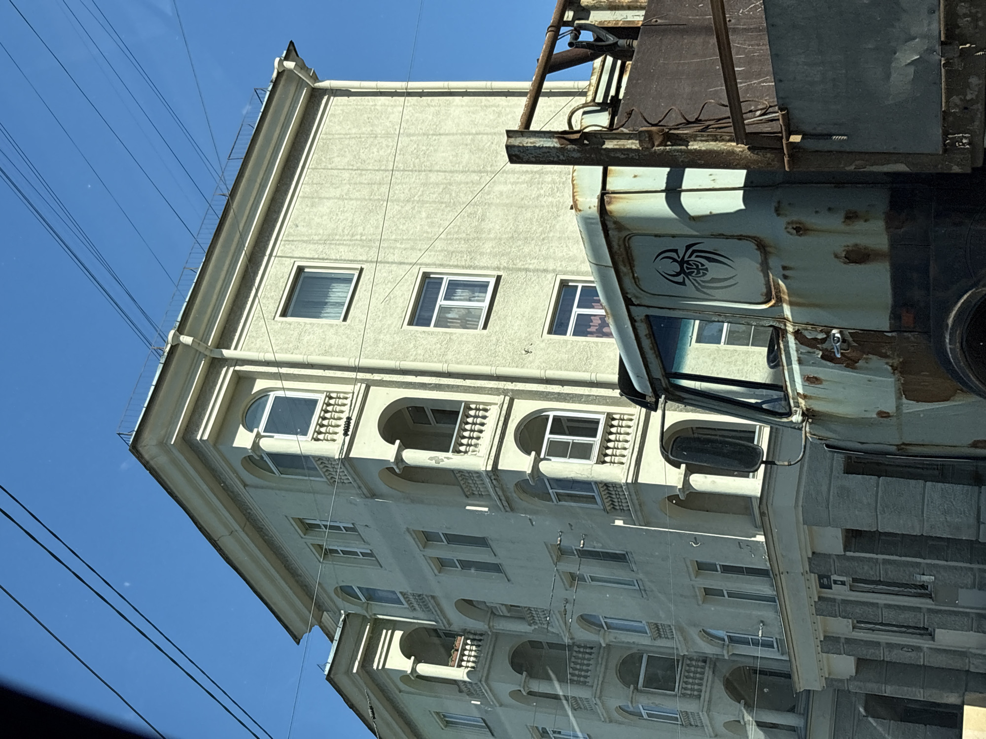 The image shows a close-up of the side of a rusty, light blue truck with a black spider logo on the door. The truck is parked in front of a pale yellow, multi-story building with arched balconies and numerous windows. The building has a classical architectural style. Several power lines are visible against a clear blue sky. The scene appears to be an urban environment, possibly in the city of Saint Petersburg, Russia, given the architectural style of the building and the presence of overhead power lines, which are common in older districts of the city. There are no people visible in the image. The truck is the most prominent object in the foreground, its weathered state contrasting with the cleaner facade of the building behind it.