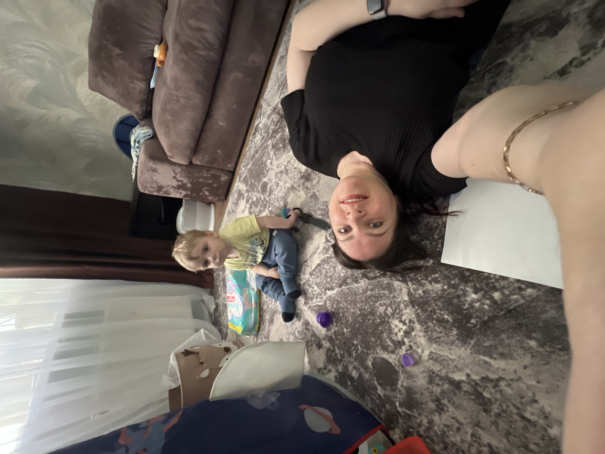 A woman and a toddler are lying on a patterned rug in a room. The woman has dark hair and is wearing a black shirt. The toddler, positioned to the left of the woman, has blonde hair and wears a yellow shirt and blue pants. A pack of diapers, a child's chair, and a blue play mat with planet designs are visible. There is also a brown couch in the background and a window with sheer curtains. Based on the information provided, the location is in Noviy, Russia.