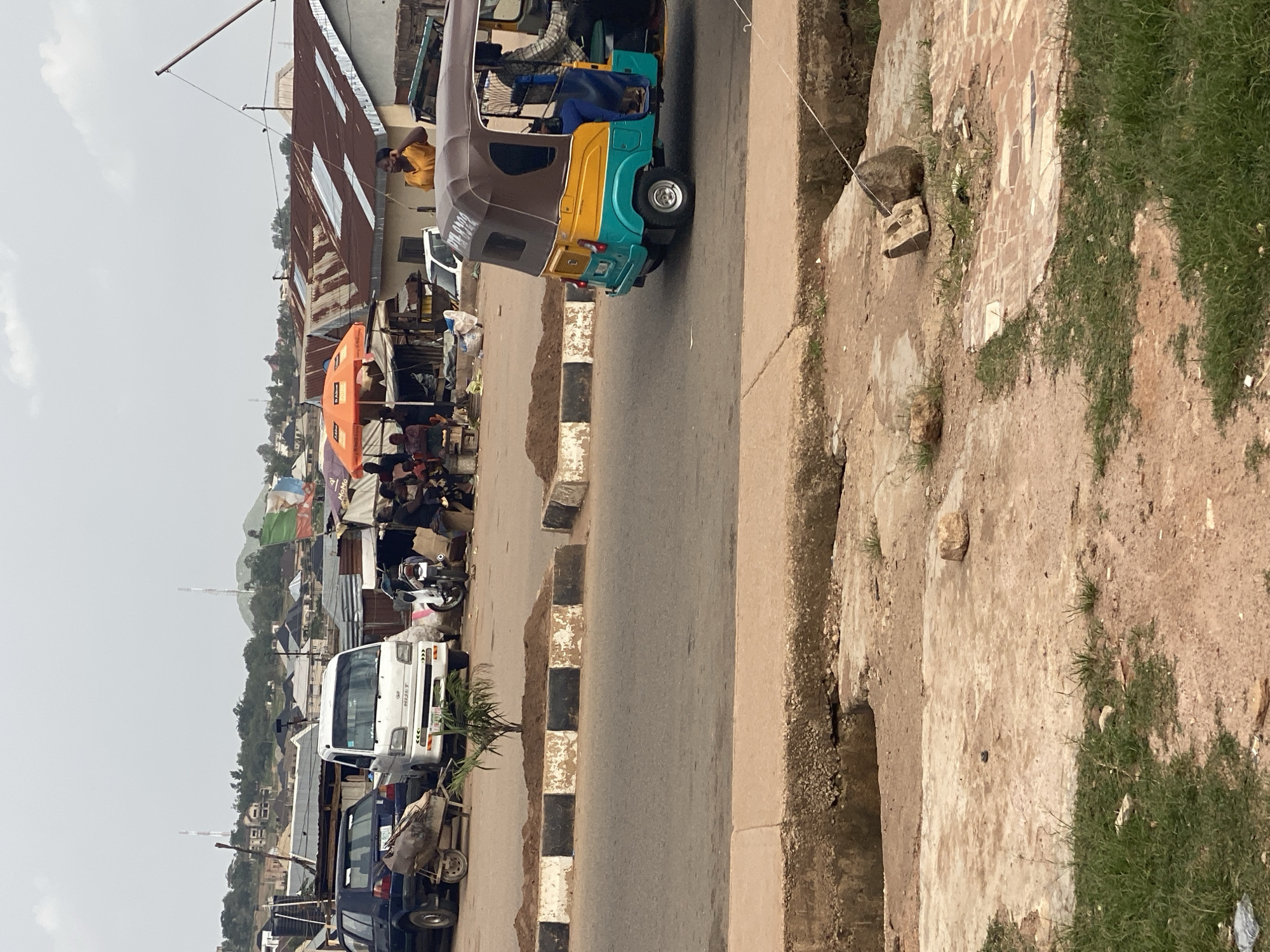 The image depicts a street scene in Gura Top, Nigeria, with a focus on the foreground and midground. A light-colored road occupies the majority of the lower portion of the frame, bordered by a concrete curb and a ditch. To the left, a patch of green grass and dirt is visible. In the midground, a busy market or roadside stalls are set up. Several people are seen in the market area, some standing and interacting. Various vehicles are present, including a white van with "HIJET" branding and a dark-colored car. A three-wheeled auto-rickshaw, commonly known as a "Keke," is driving on the road in the right midground, with two individuals inside. Behind the stalls, buildings with corrugated metal roofs are visible, indicating a developing urban or semi-urban environment. In the background, a hilly landscape with more buildings and communication towers suggests the broader Gura Top setting. The sky is overcast, contributing to the diffused lighting of the scene.