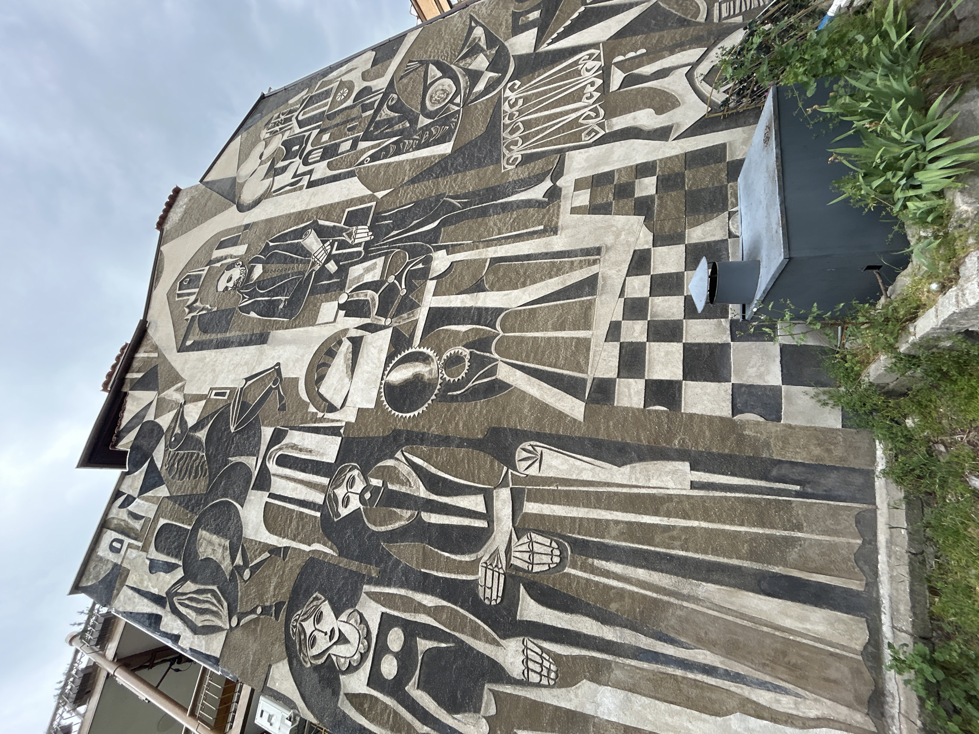 The image depicts a large, monochromatic mural on the side of a building in Plovdiv, Bulgaria. The mural is rendered in shades of grey, black, and beige and features a stylized, abstract depiction of people and objects. The figures are elongated and fragmented, with elements resembling cubist or futurist art. The mural includes images of faces, hands, and various geometric shapes. A section of the mural transitions into a checkered pattern. A grey metal utility box is attached to the building near the mural. Green vegetation grows along the base of the building. The sky above the building is overcast.