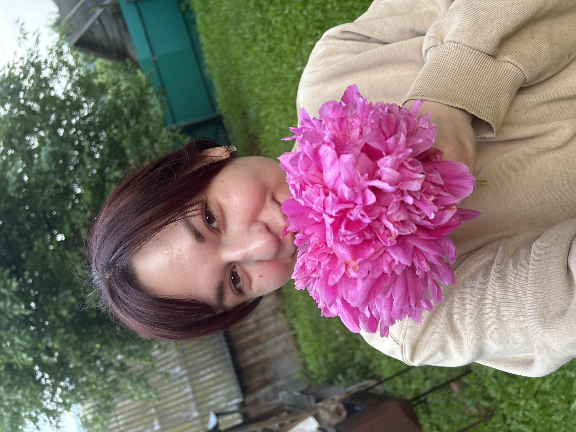 The image shows a young woman with dark hair holding a large, vibrant pink peony flower up to her face. She is wearing a beige sweatshirt. She is in an outdoor setting, likely a backyard, with green grass and trees visible in the background. A wooden fence and a rustic building are also present, suggesting a rural or suburban environment. The overall scene is casual and outdoorsy.