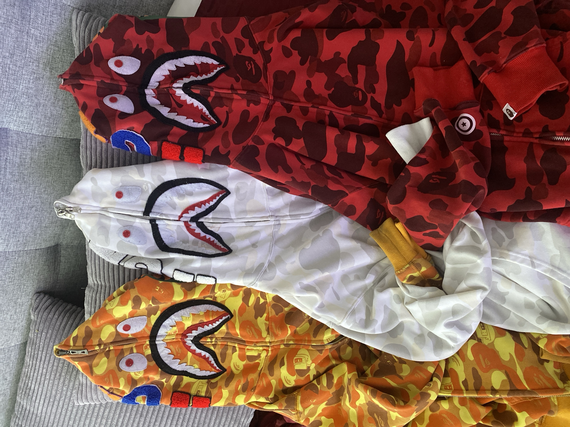The image features three BAPE (A Bathing Ape) "shark" full-zip hoodies, laid out on a textured grey surface, likely a ribbed sofa or blanket, within an indoor setting. The hoodies are arranged diagonally from the top-left to the bottom-right, slightly overlapping.
The top hoodie is red with a camouflage pattern in varying shades of red. Its hood forms a shark face when zipped up, displaying white teeth with black outlines, prominent red "eyes" with black pupils, and a blue and red patch on the left side of the "mouth." A small white tag with a black logo, characteristic of the BAPE brand, is visible on the right cuff.
The middle hoodie is white and light grey, also in a camouflage pattern. Its hood similarly features white shark teeth with black outlines and red "eyes" with black pupils.
The bottom hoodie is an orange, yellow, and brown camouflage design. Its hood mirrors the others with white shark teeth, black outlines, red "eyes" with black pupils, and blue and red patches on the left side of the "mouth."
All hoodies showcase the distinctive full-zip design that extends through the hood, creating the iconic shark face. The fabric appears thick and soft. The grey background has a distinct, consistent ribbed pattern. The items are neatly arranged, suggesting they are being displayed or stored. No human activity is depicted. No discernible text is visible beyond the brand's logo on one cuff. The image itself provides no visual cues to confirm the specified location of Sochi, Russia.