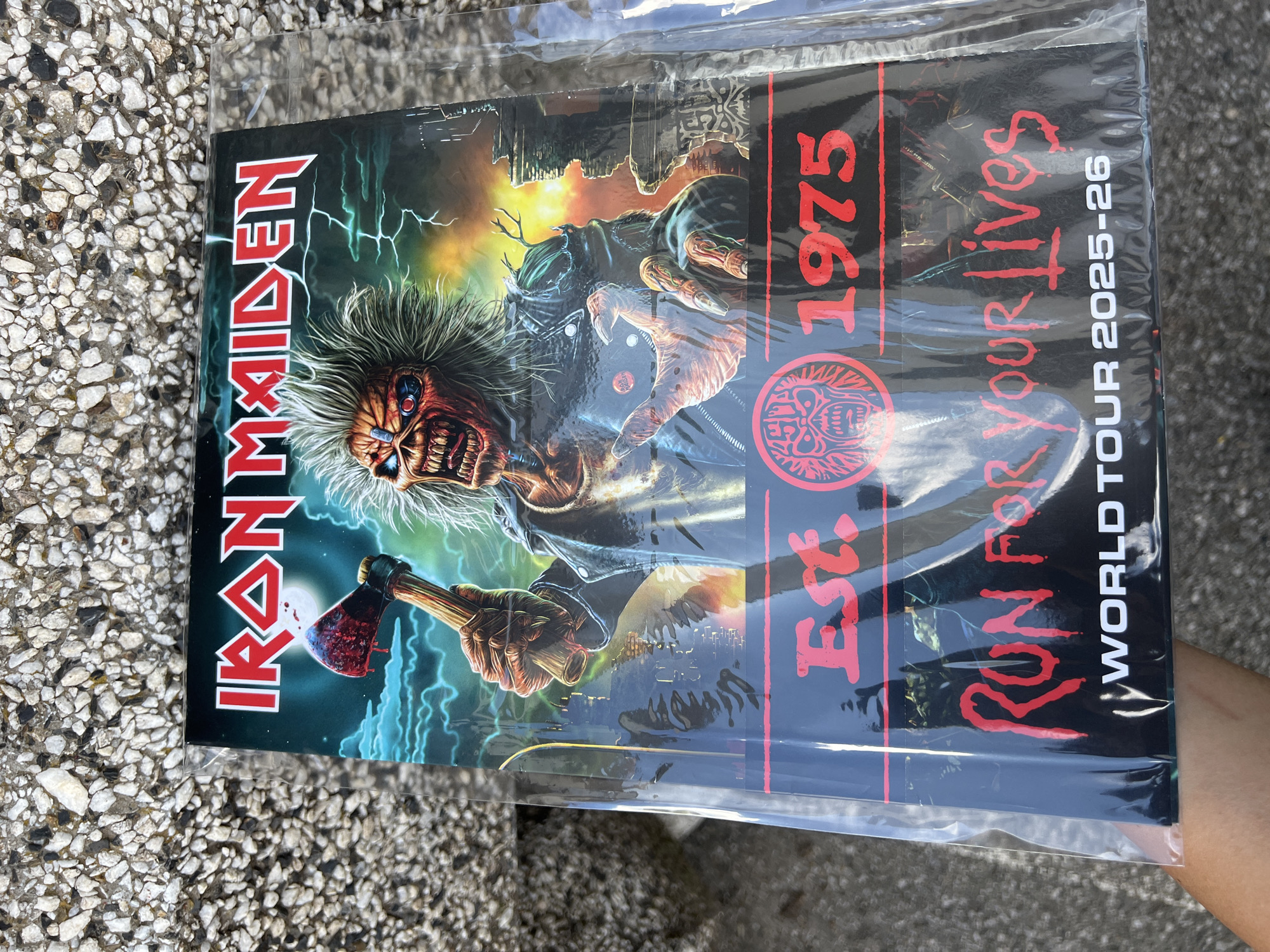 A poster featuring the Iron Maiden mascot Eddie, holding a bloody axe, with the words "IRON MAIDEN", "Est. 1975", and "RUN FOR YOUR LIVES WORLD TOUR 2025-26" is laying on an outdoor paved surface. The poster is in a plastic sleeve. The paved surface appears to be light-colored with small, dark pebbles. There is no indication of the city of Sofia, Bulgaria.